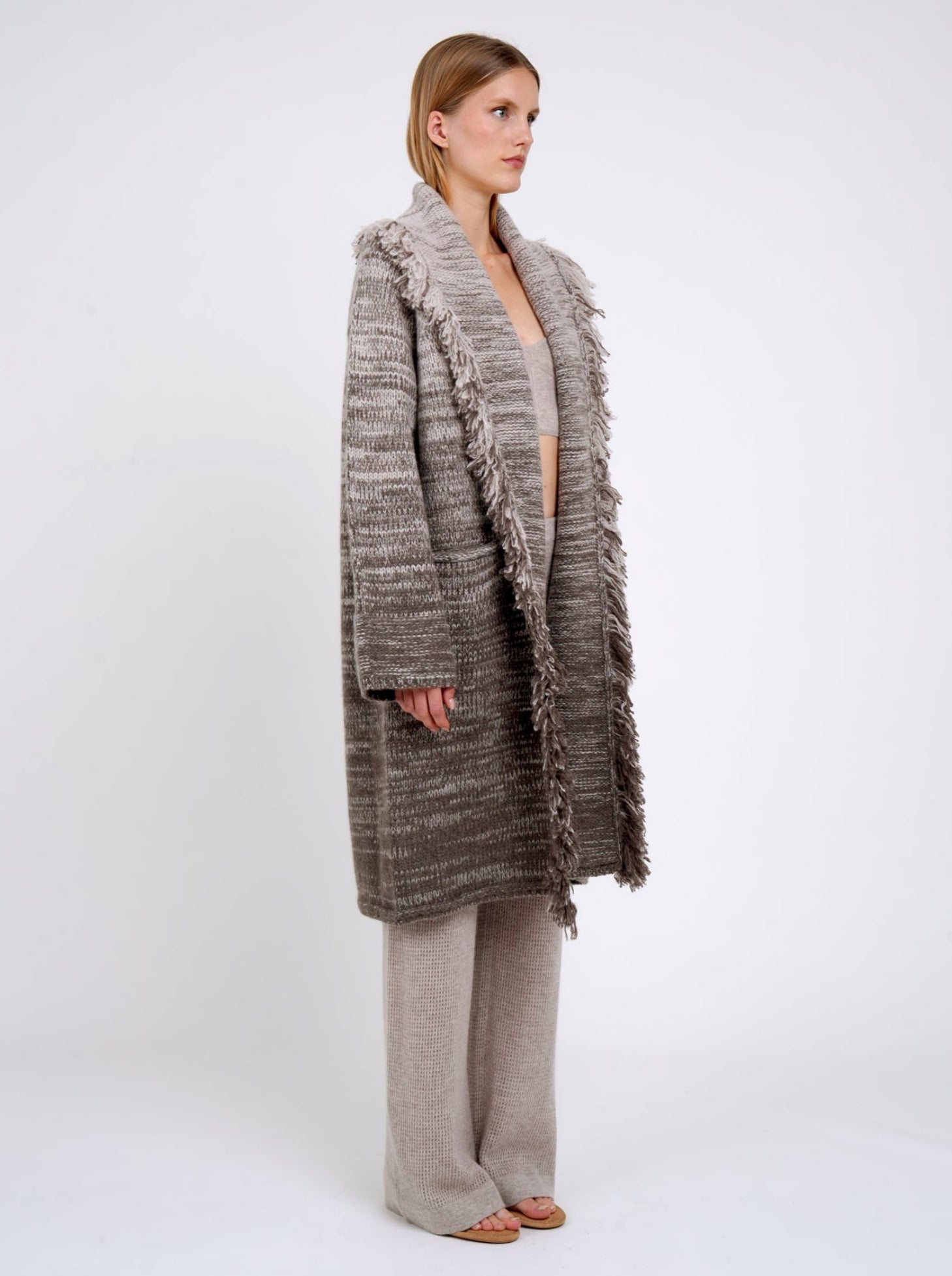 Side view of model in mixed colour knitted coat in beige and brown with fringed shawl collar, long wide sleeves; with beige cashmere knit pants.