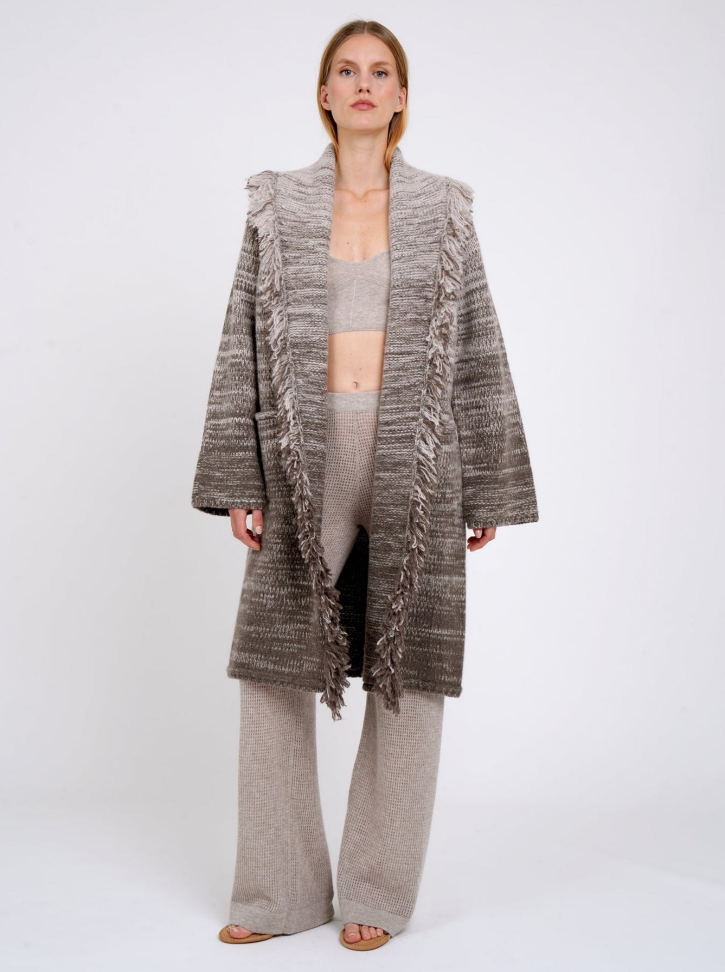 Model wears mixed colour knitted coat in beige and brown with fringed shawl collar, long wide sleeves; worn with beige cashmere knit pants and beige knit cropped bralette top.