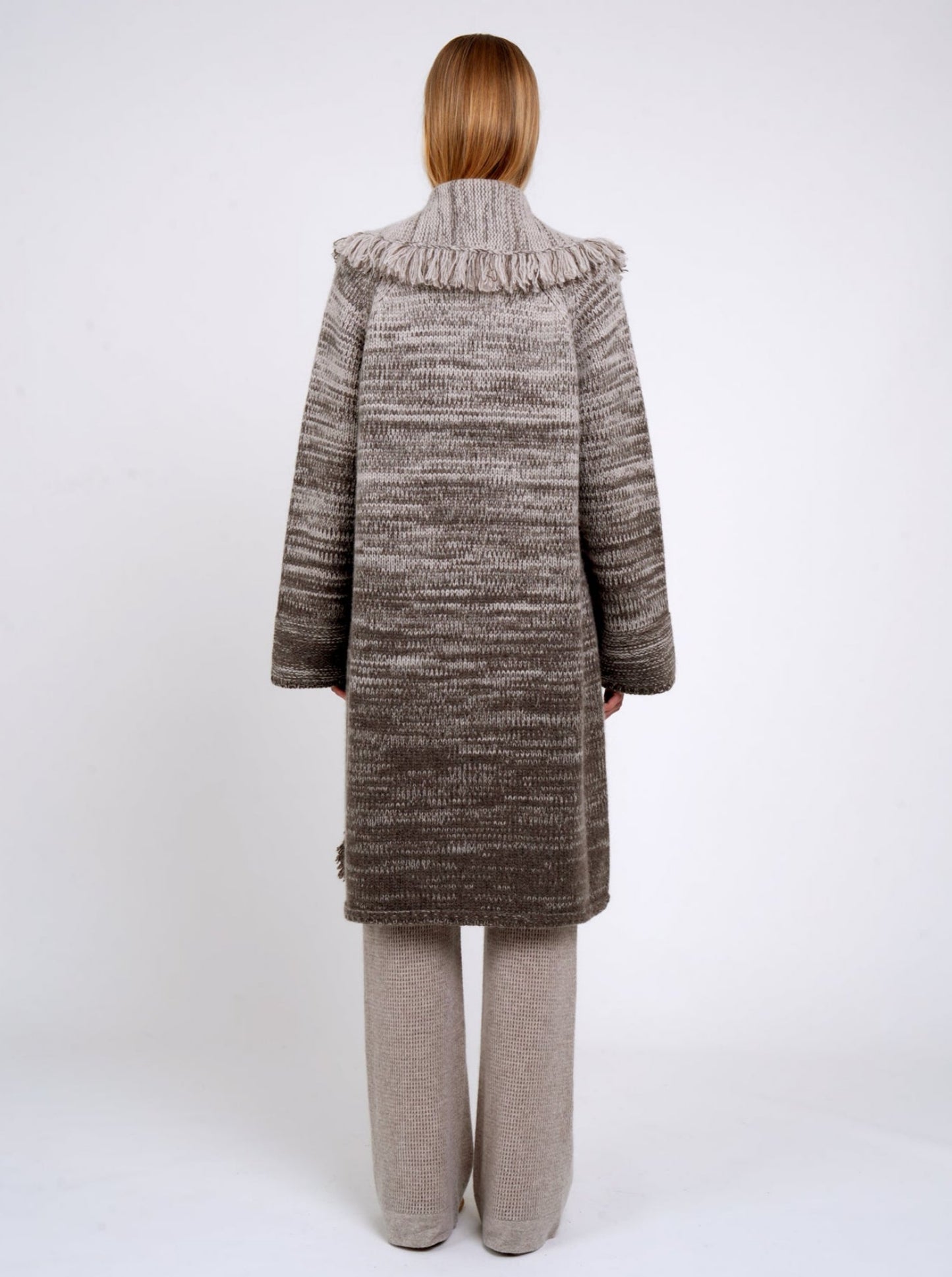 Back view model wearing mixed colour knitted coat in beige and brown with fringed shawl collar, long wide sleeves; with beige cashmere knit pants.