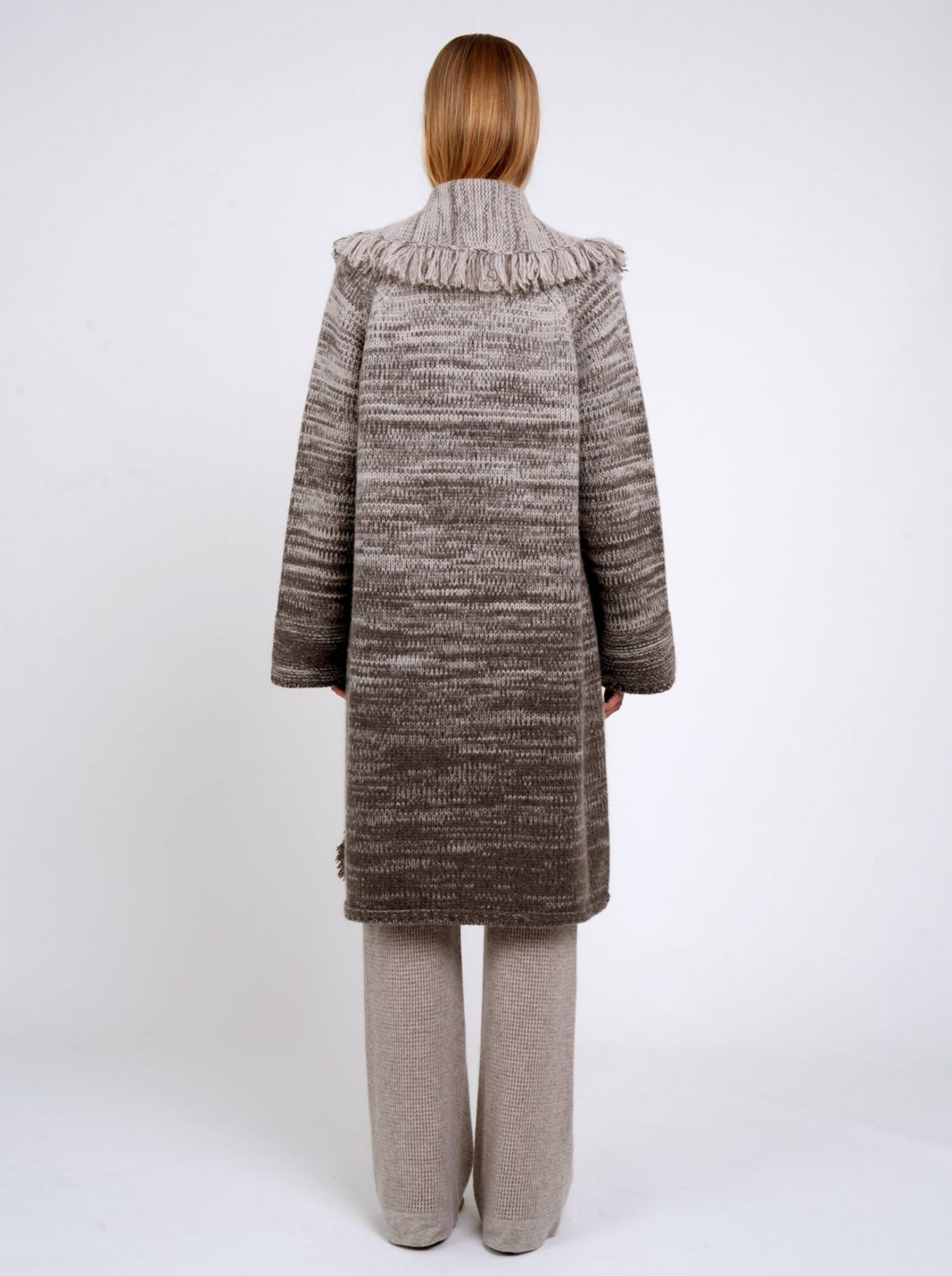 Back view model wearing mixed colour knitted coat in beige and brown with fringed shawl collar, long wide sleeves; with beige cashmere knit pants.