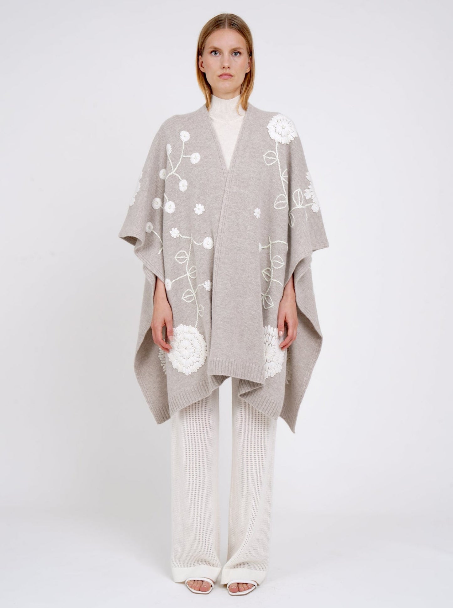 front view model in beige cashmere long shawl with large white embroidered flowers over white knit  turtle neck top and white pants.