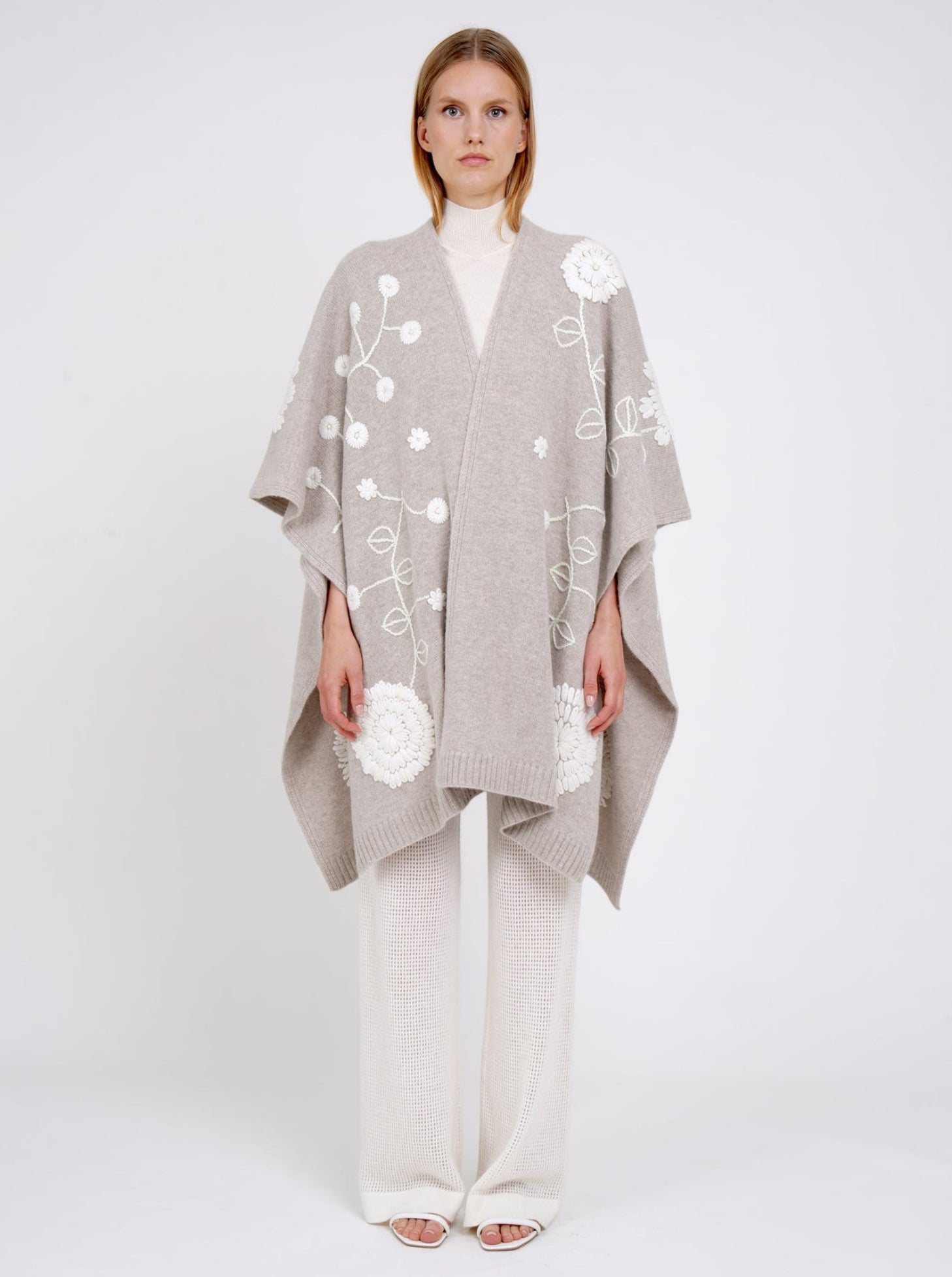 front view model in beige cashmere long shawl with large white embroidered flowers over white knit  turtle neck top and white pants.
