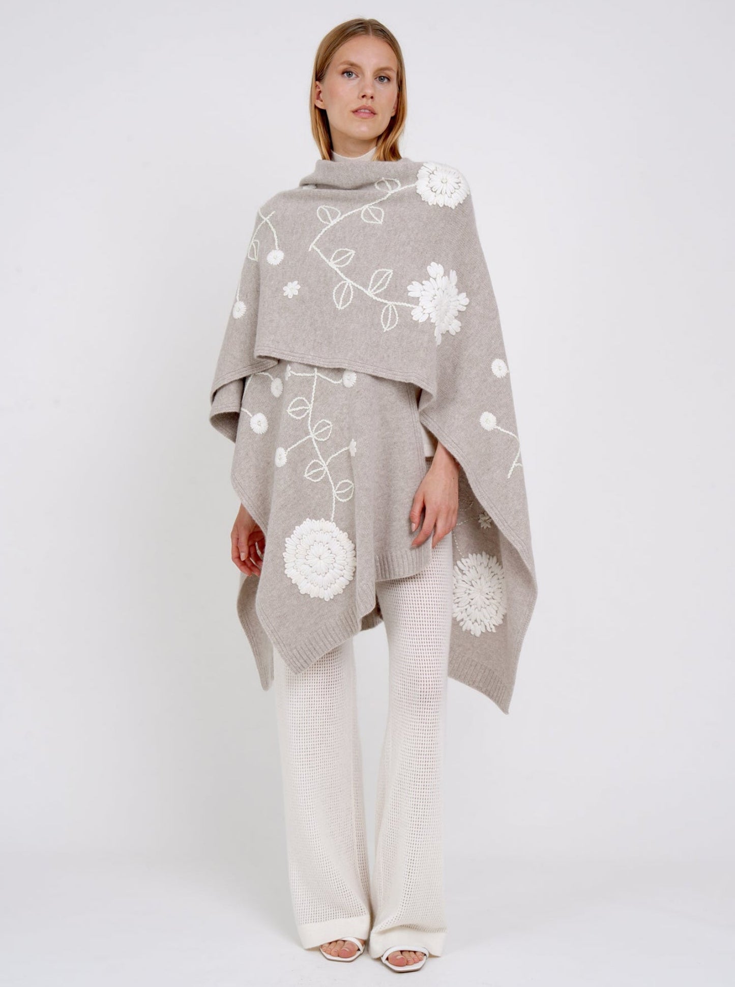 model wears beige cashmere long shawl with large white embroidered flowers over white knit pants.