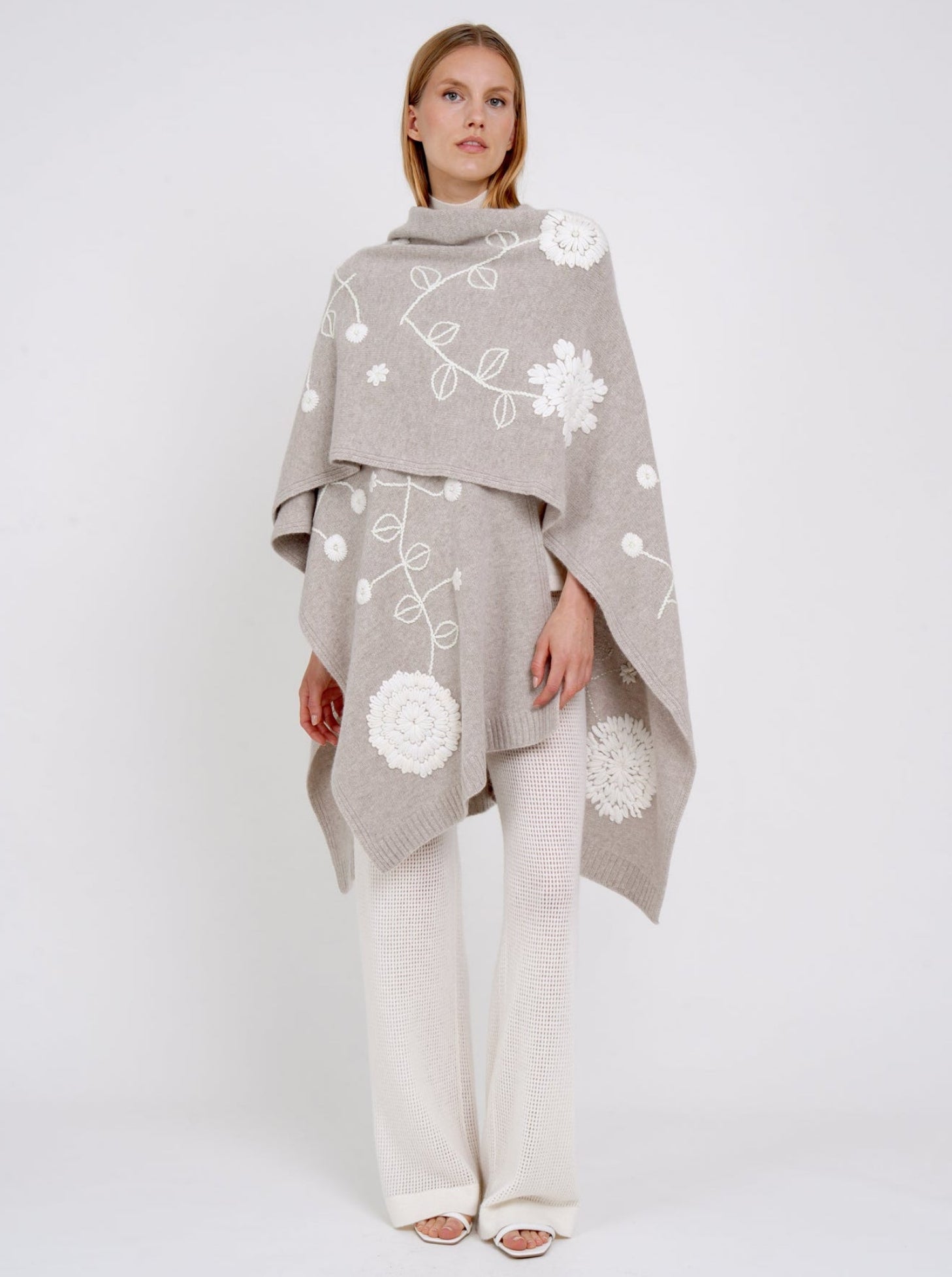 model wears beige cashmere long shawl with large white embroidered flowers over white knit pants.