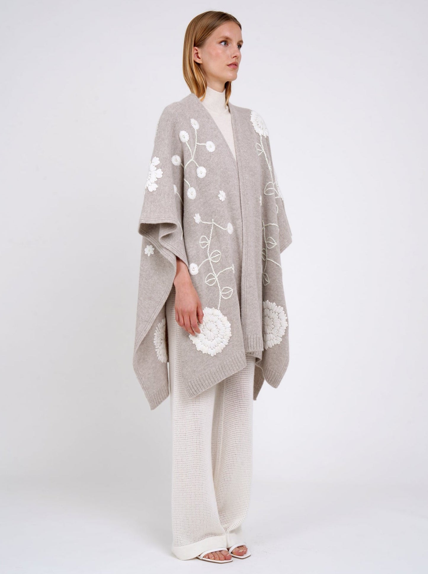 Side view model in beige cashmere long shawl with large white embroidered flowers over white knit  turtle neck top and white pants.