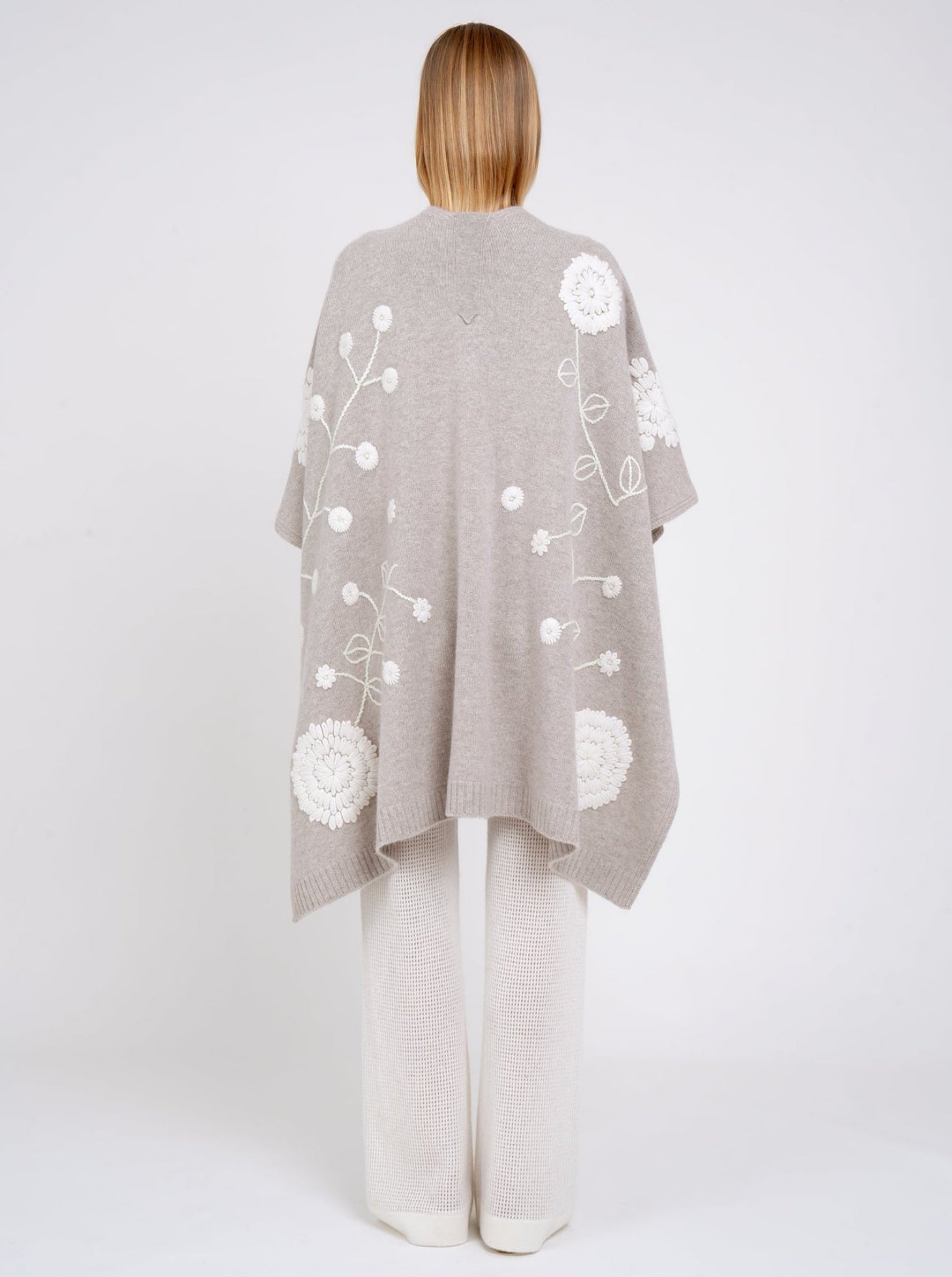 Back view model in beige cashmere long shawl with large white embroidered flowers over white knit pants.
