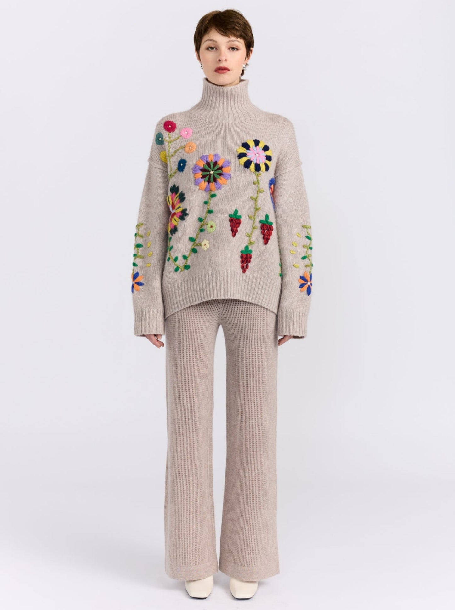 Model wearing light beige oversized roll neck jumper adorned with brightly coloured hand embroidered flowers; worn with beige net knit wide leg pants.