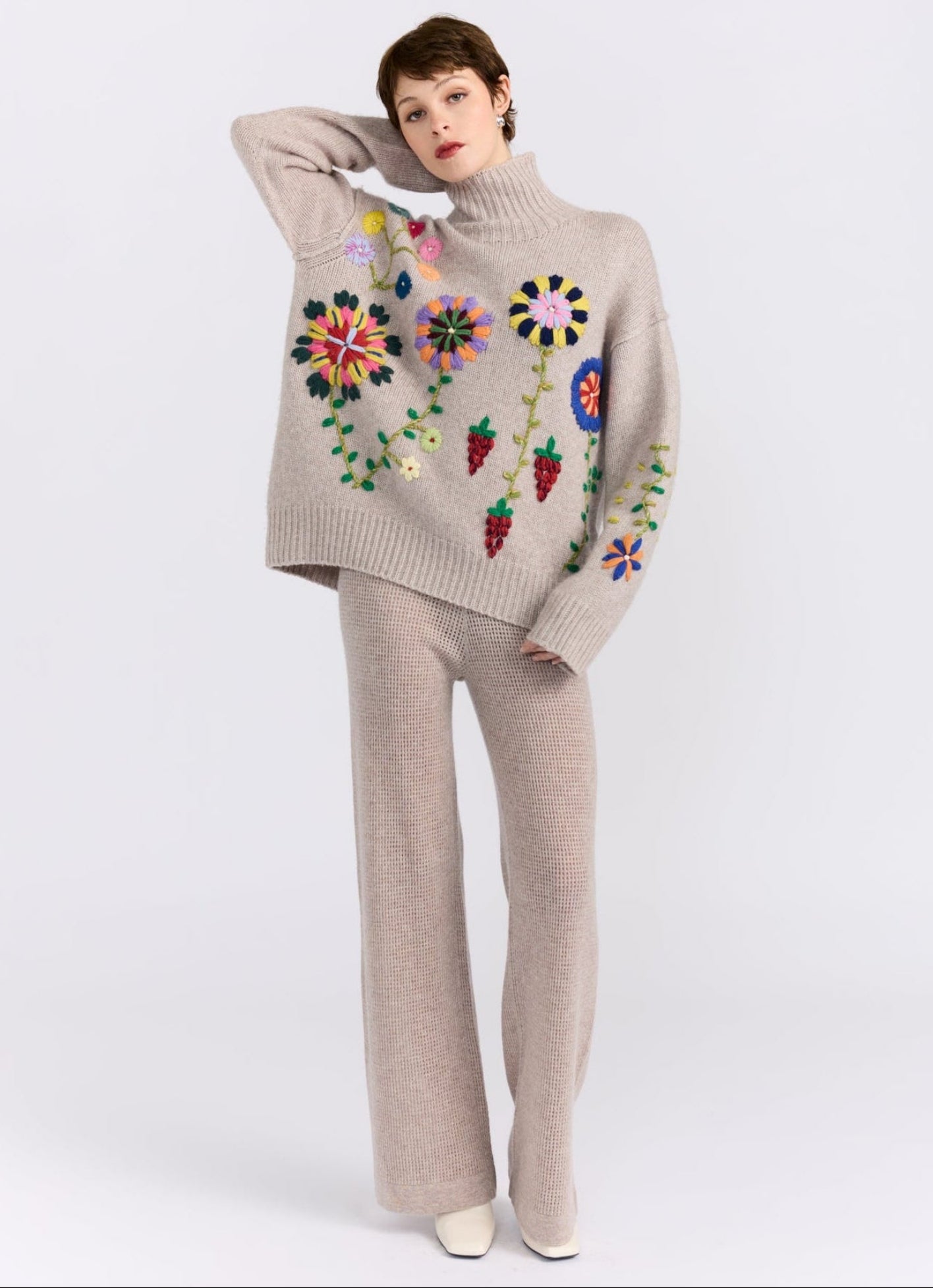 Model poses in light beige  chunky knit oversized roll neck jumper with brightly coloured hand embroidered flowers; worn with matching beige net knit straight leg pants.