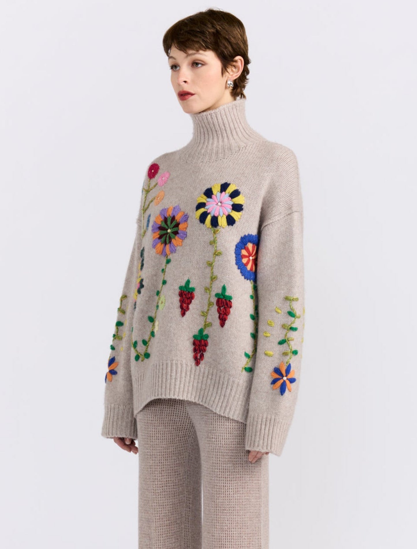 Cropped image side view model in chunky knit beige oversized roll neck jumper with brightly coloured hand embroidered flowers; and beige net knit straight leg pants.