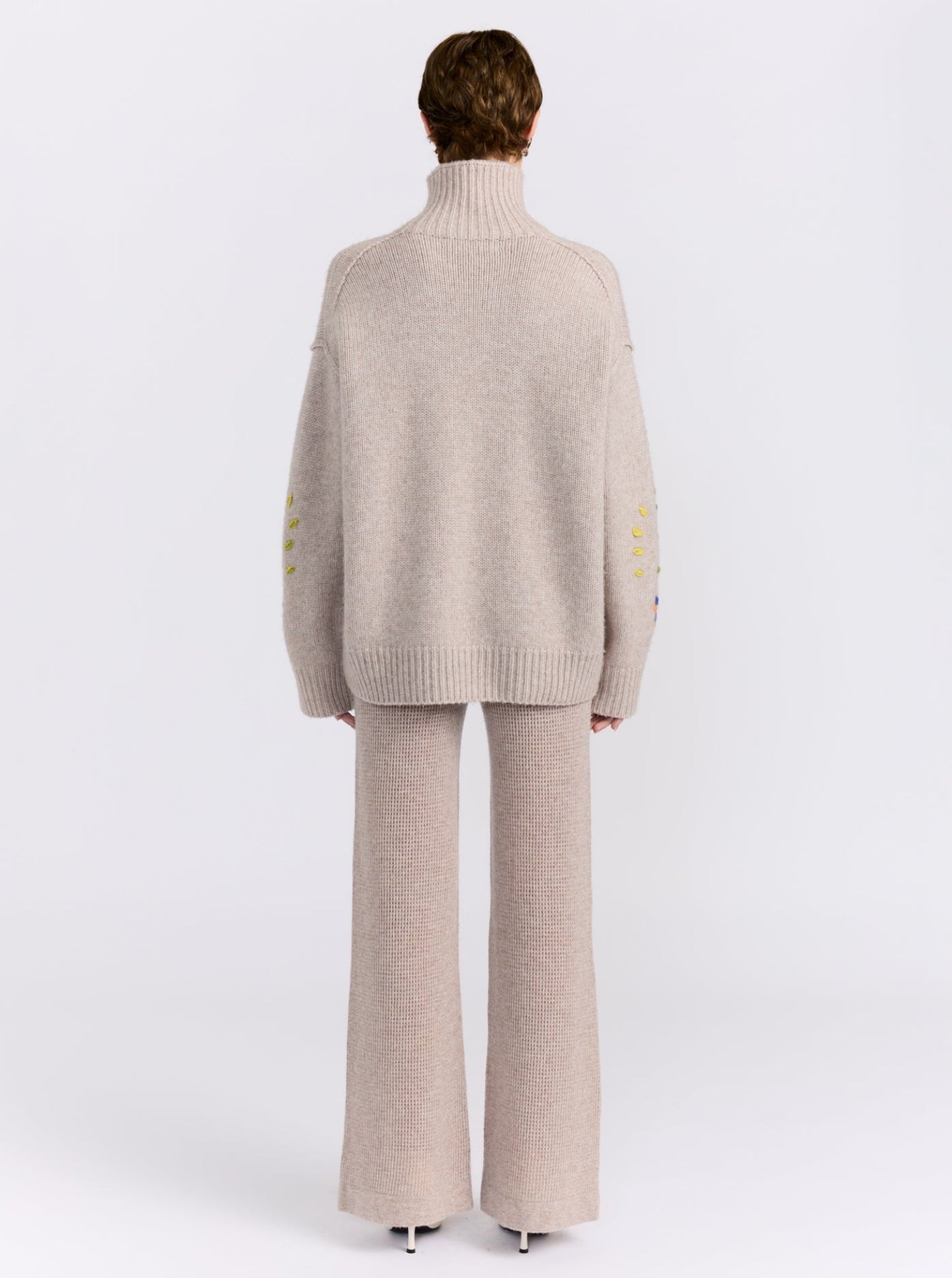 Back view model wearing chunky knit beige oversized roll neck jumper; worn with beige net knit wide leg pants.