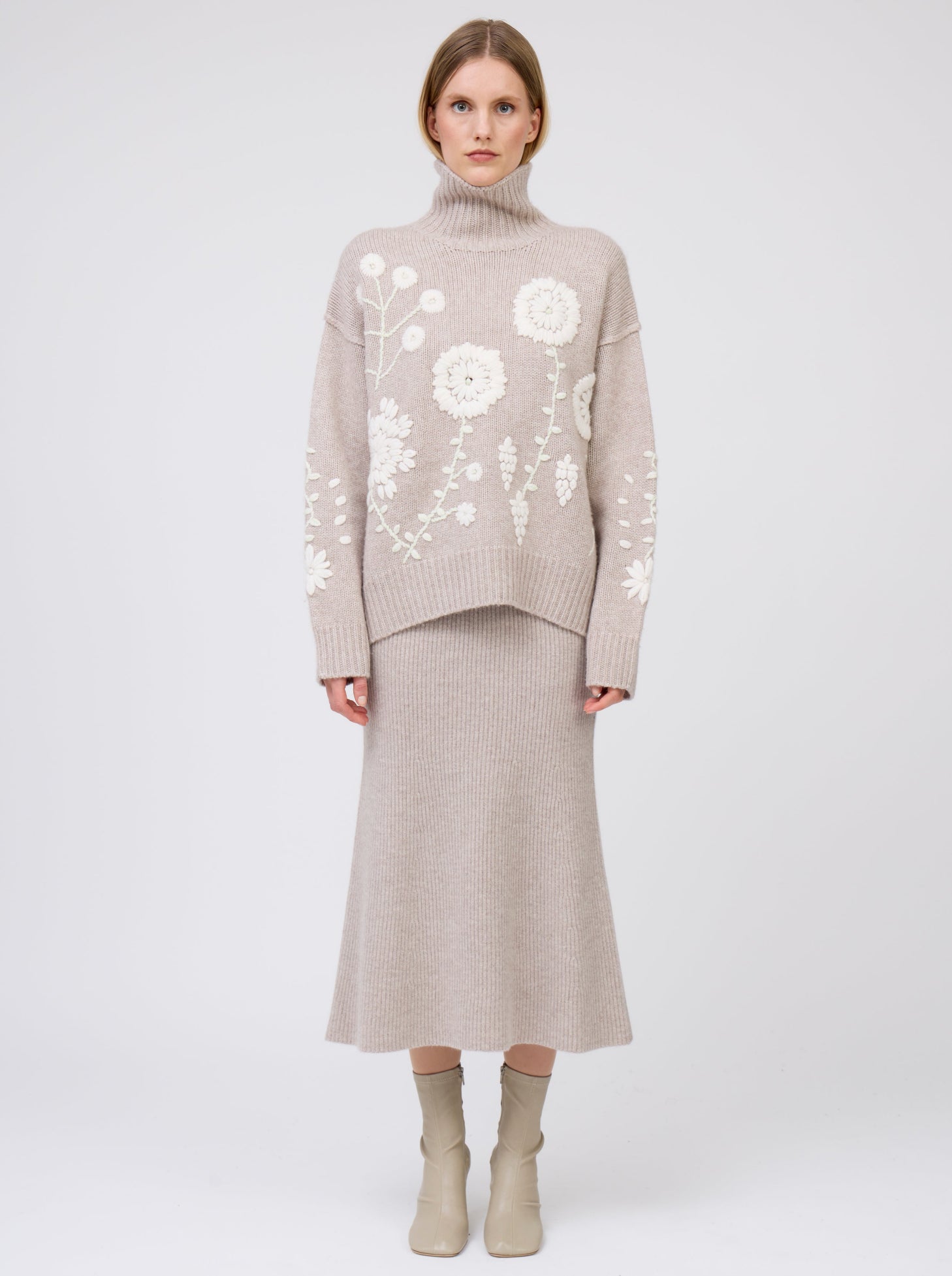 model wears chunky knit beige sweater with embroidered white flowers; beige knit flared skirt & beige ankle boots.