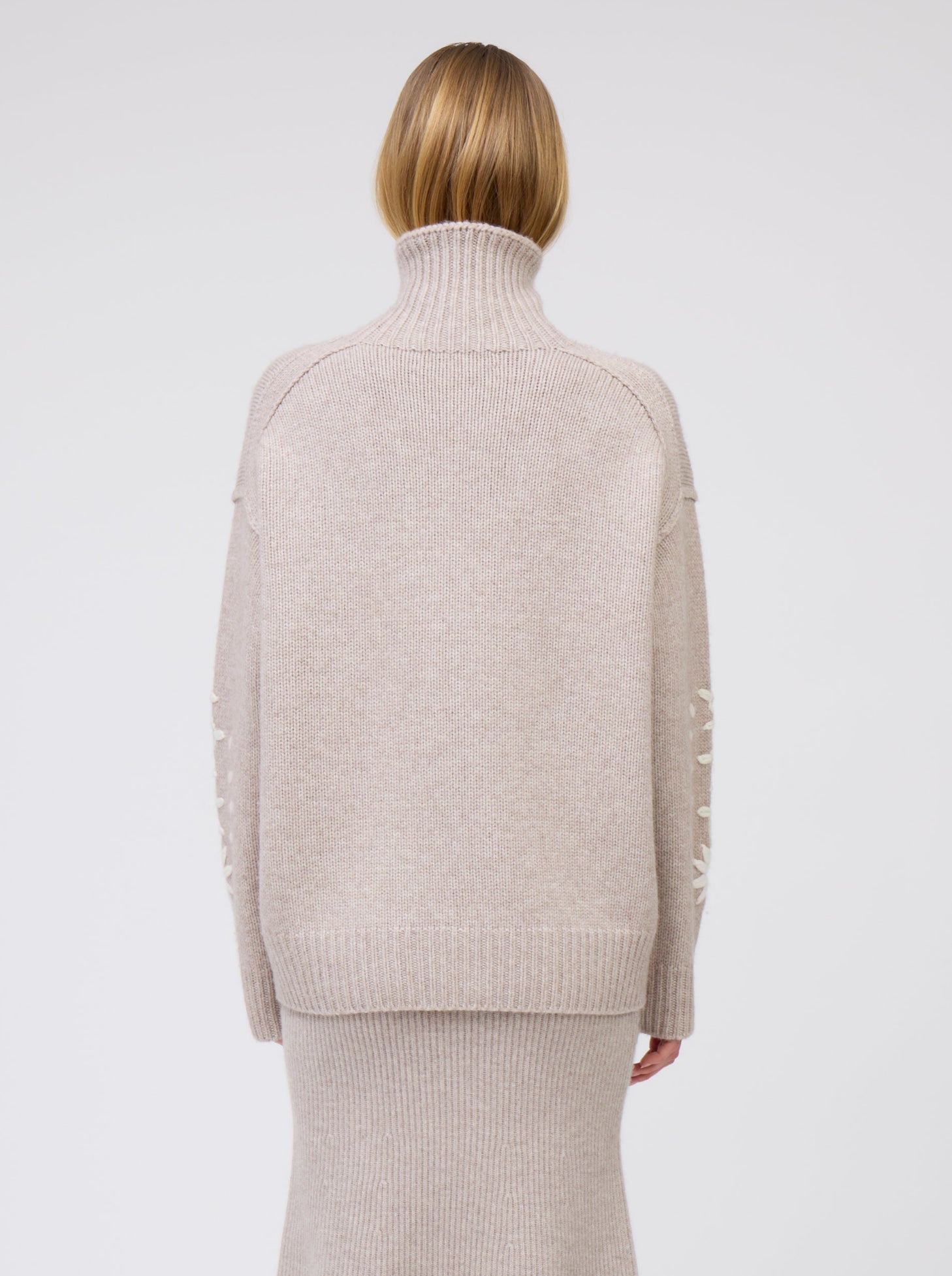 Back view model in chunky knit beige sweater with high turtle neck and beige knit flared skirt.