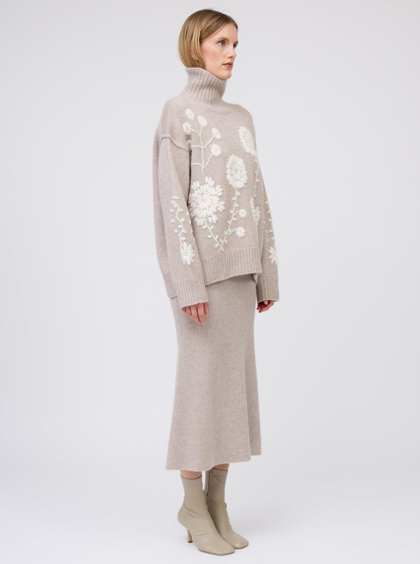 Side view model wearing chunky knit beige roll neck sweater with embroidered white flowers; beige knit flared skirt & beige ankle boots.