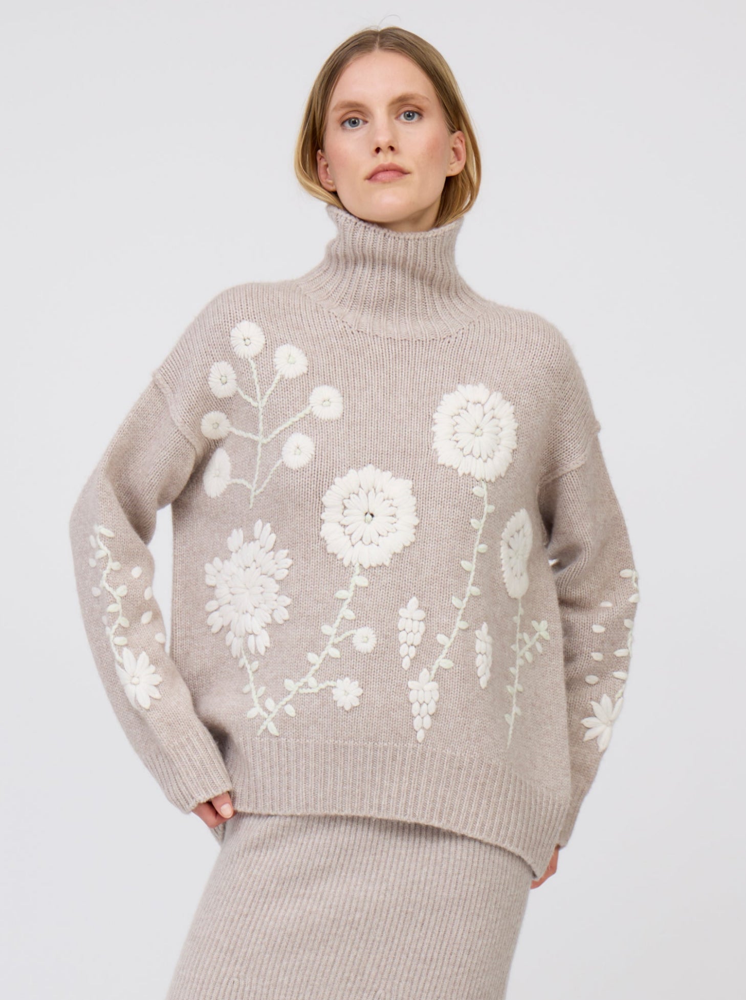Close up of model in chunky knit beige sweater with embroidered white flowers and high turtle neck.