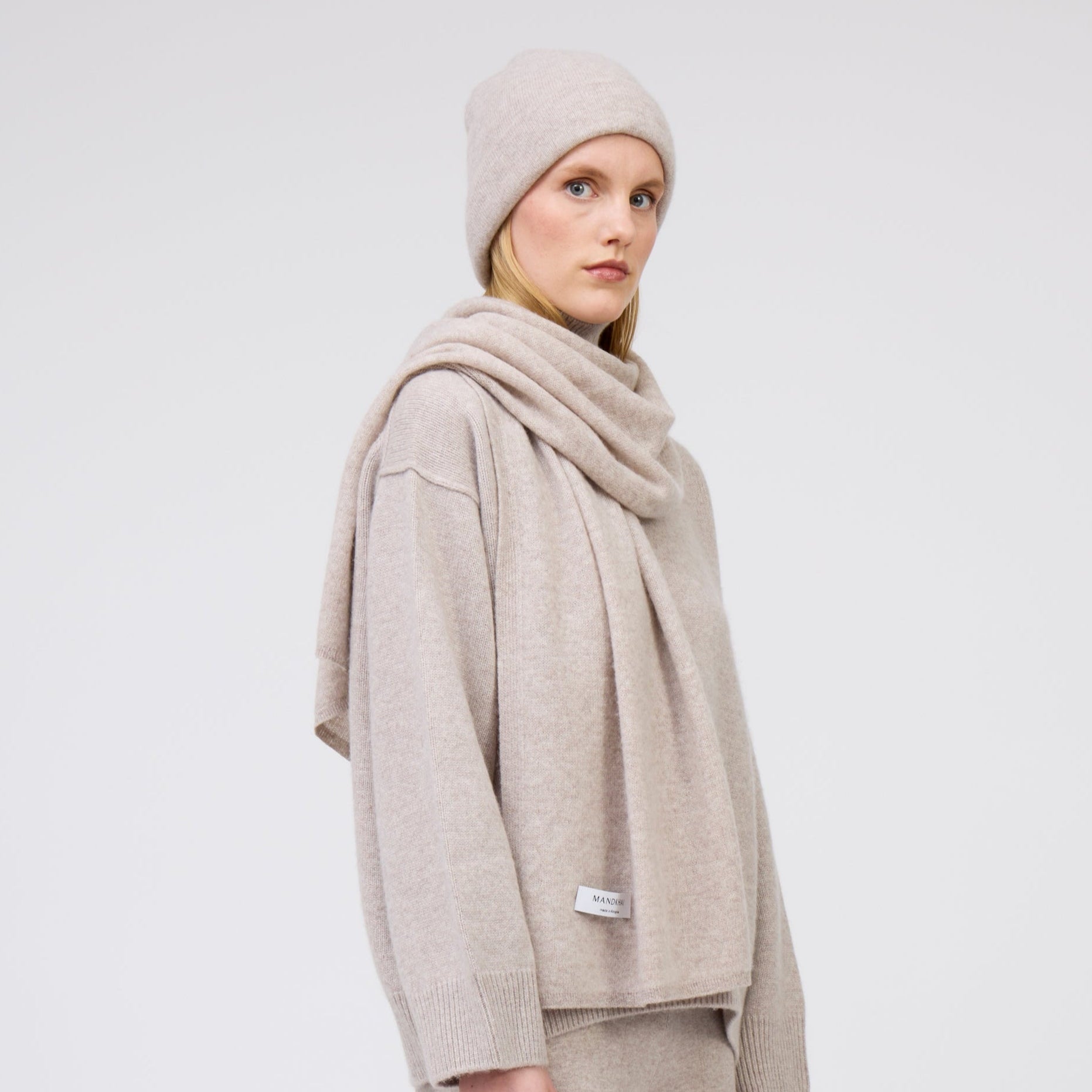 Cropped side view of model wearing complete outfit in matching beige cashmere: beanie hat, scarf and oversized jumper.
