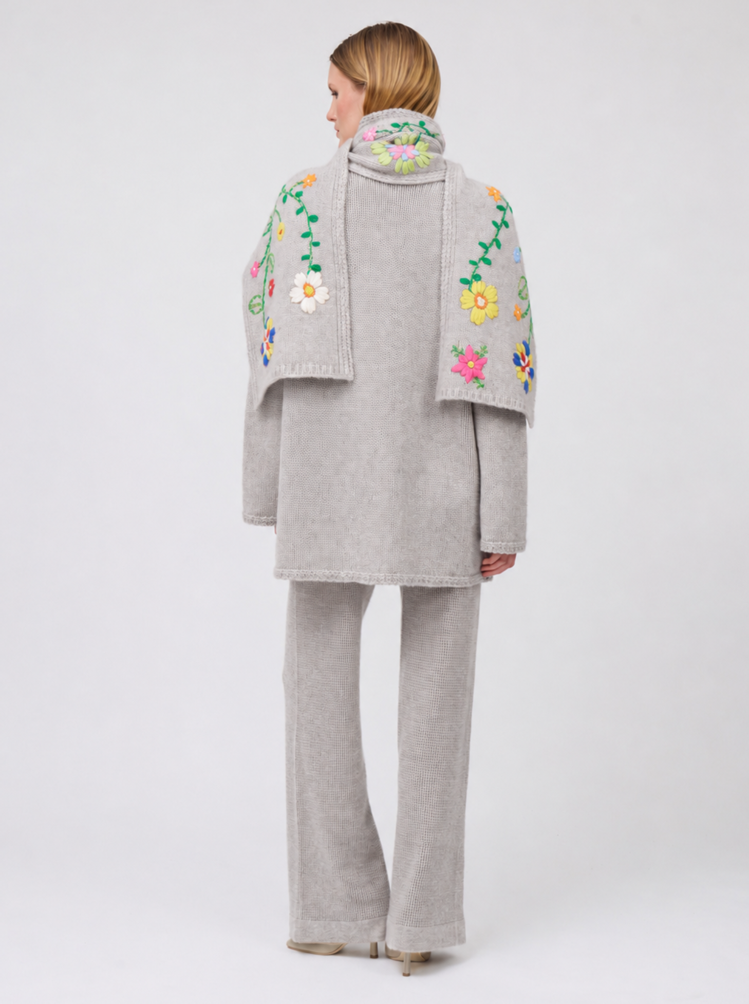 Back view model in pale beige knitted jacket with flower embroidered scarf and matching cashmere knit pants.