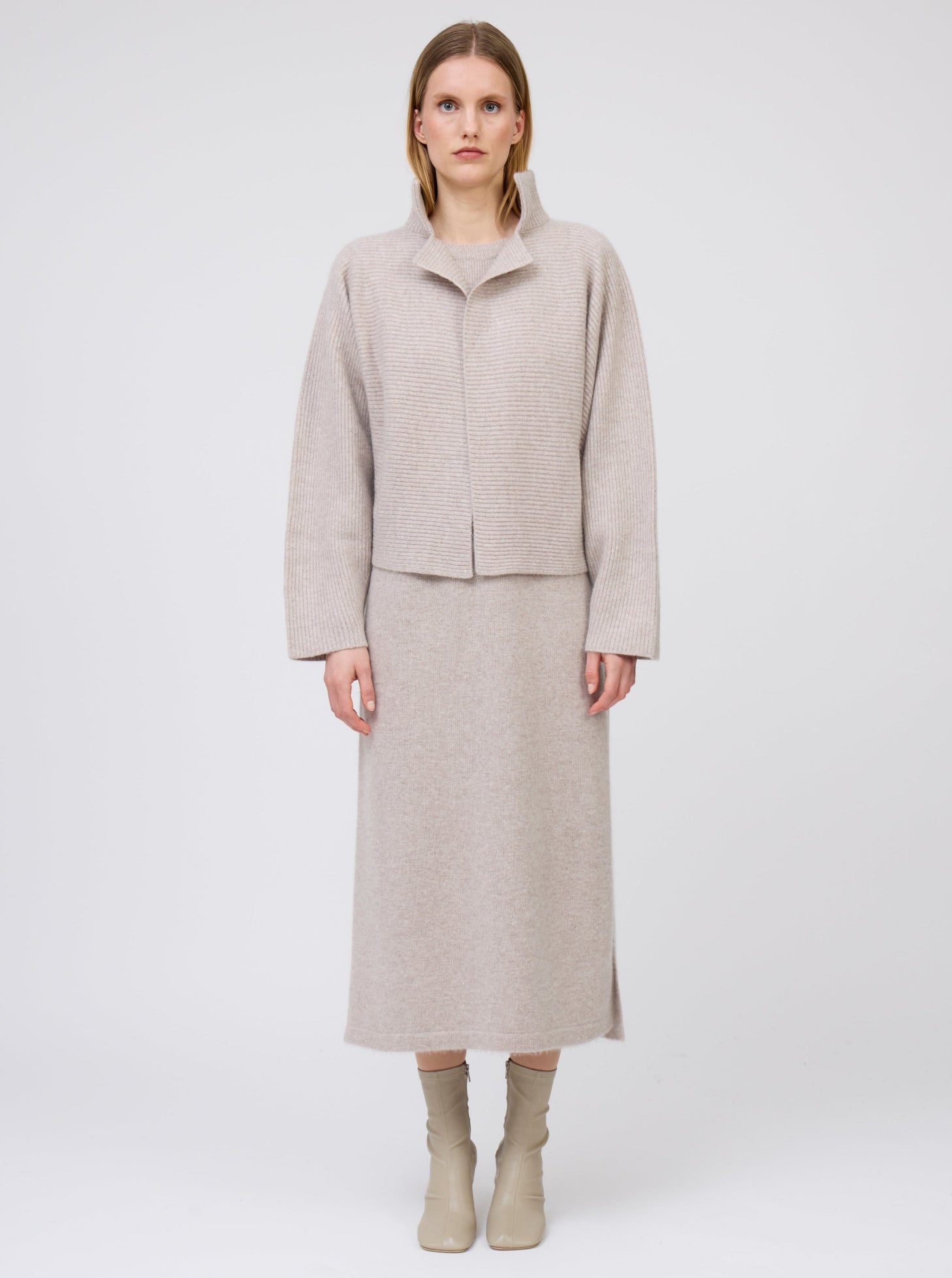 Front view of model wearing beige cashmere, ribbed knit, short jacket with high collar and kimono sleeves over matching beige cashmere straight cut dress and beige boots.