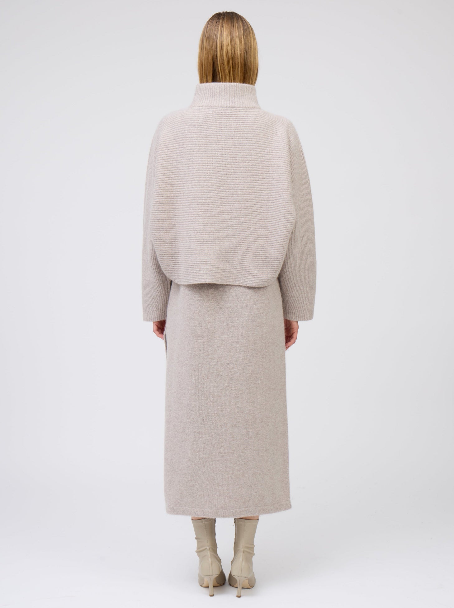 Back view of model wearing beige cashmere, ribbed knit, short kimono jacket with high collar over matching beige cashmere straight cut dress and beige boots.