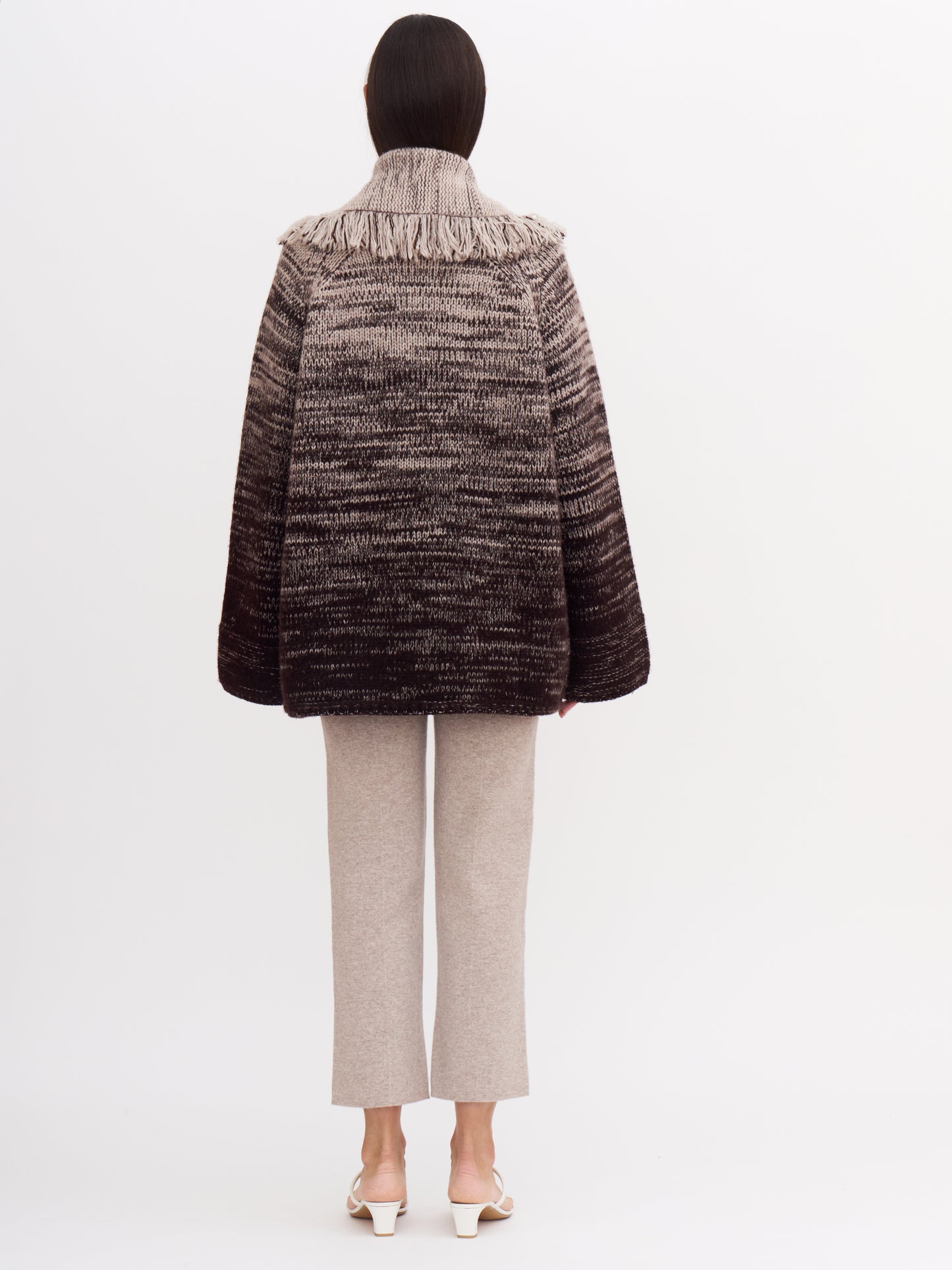 Back view of model wearing chunky knit jacket with fringed shawl collar in a cashmere colour mix of beige and dark brown; with beige cashmere cropped pants.