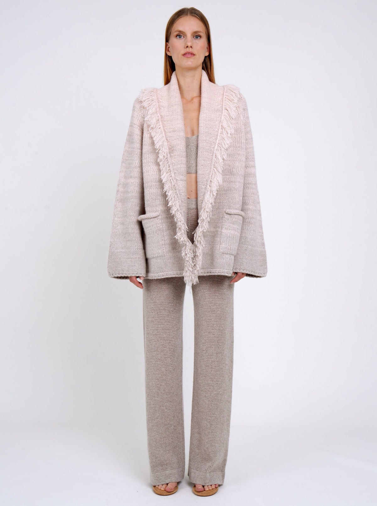 model wearing a hip-length cashmere knitted cardigan style jacket with deep shawl collar with fringes in pastel colours of beige and peach, worn with beige pants. 