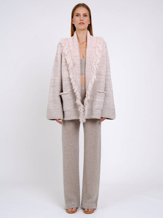 model wearing a hip-length cashmere knitted cardigan style jacket with deep shawl collar with fringes in pastel colours of beige and peach, worn with beige pants. 
