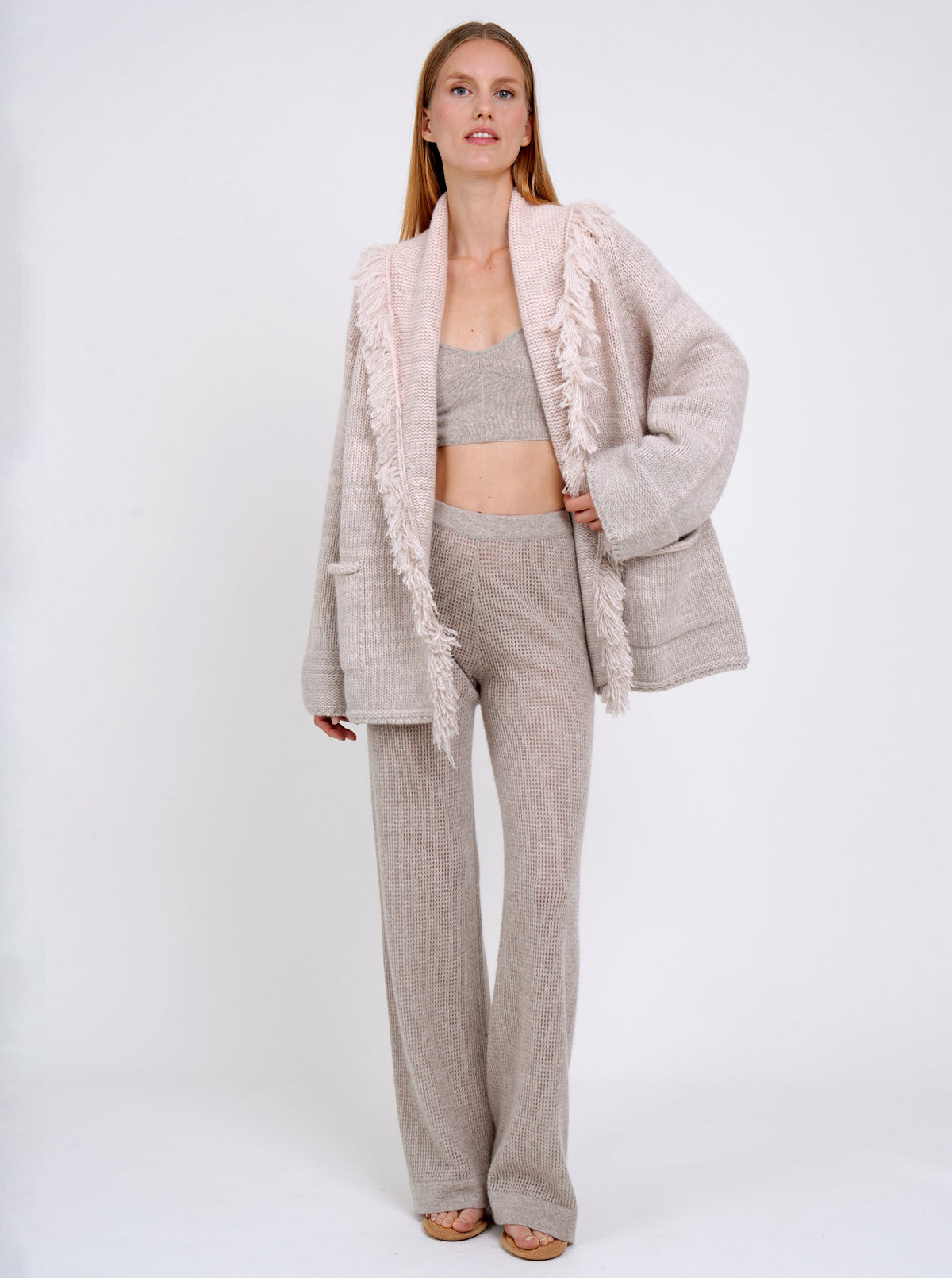 Front view model in hip-length cashmere knitted cardigan style jacket with deep shawl collar with fringes in pastel colours of beige and peach, showing beige knit bralette underneath and beige pants. 