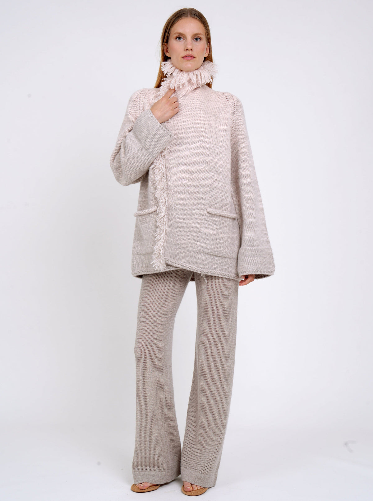model wears chunky knit cashmere jacket with fringe details in beige and peach mix colours, and high collar wrapped close and beige knit pants.
