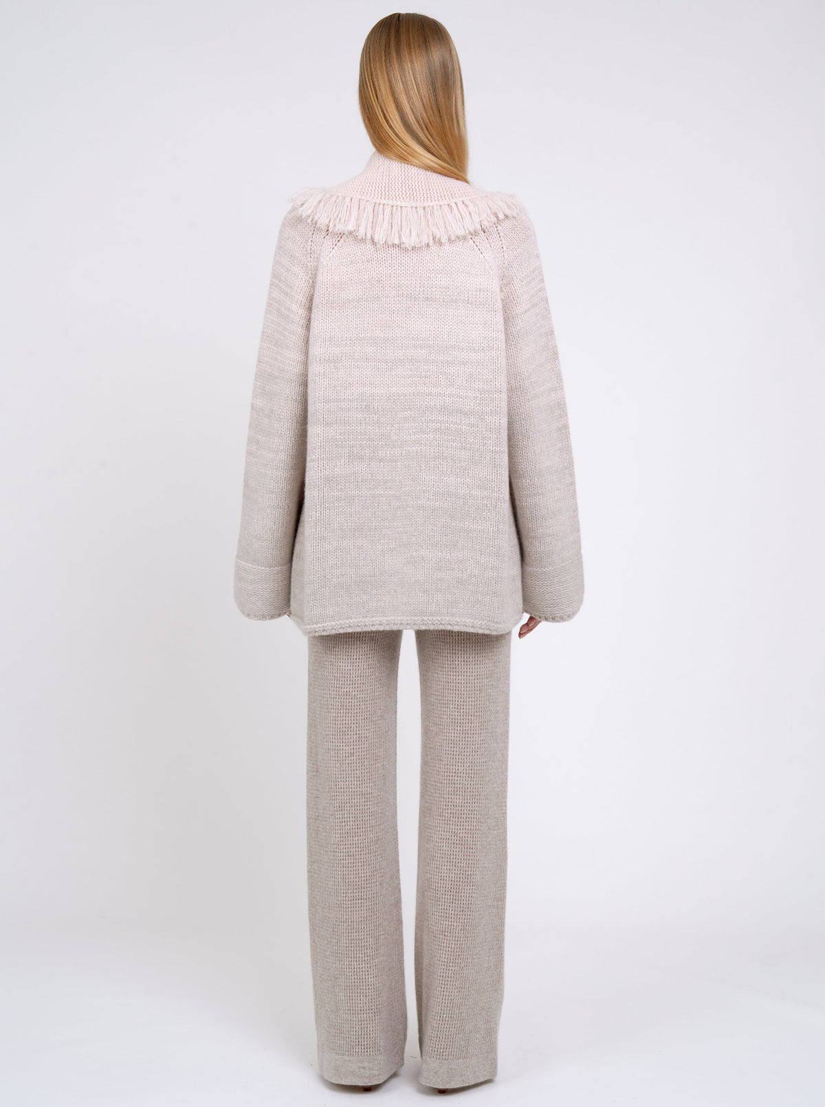 Back view model in chunky knit cashmere jacket in beige with fringed shawl collar and beige knit pants.