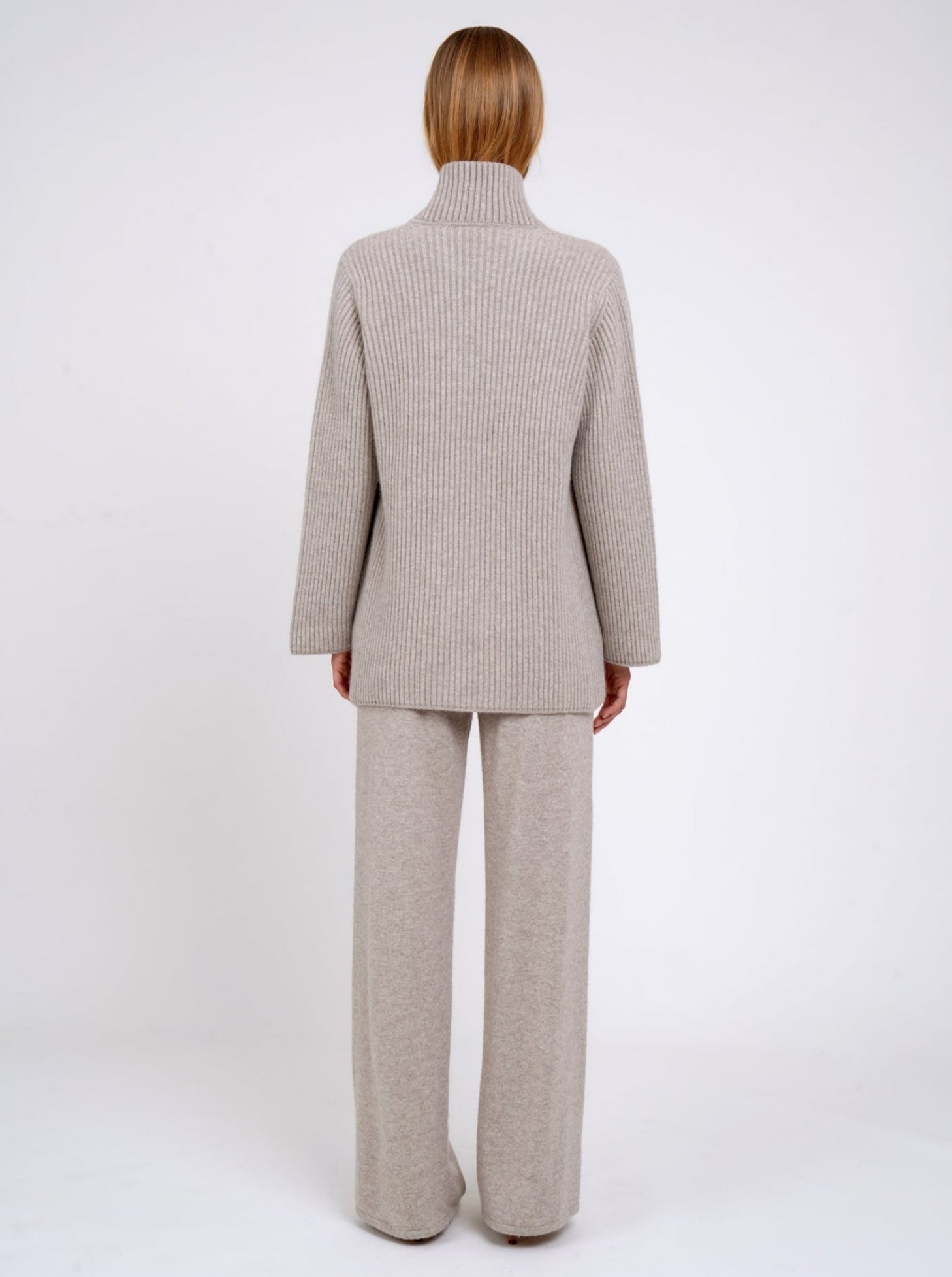 Back view model wearing beige ribbed knit cashmere jacket with high collar and patch pockets; worn with beige cashmere pants.
