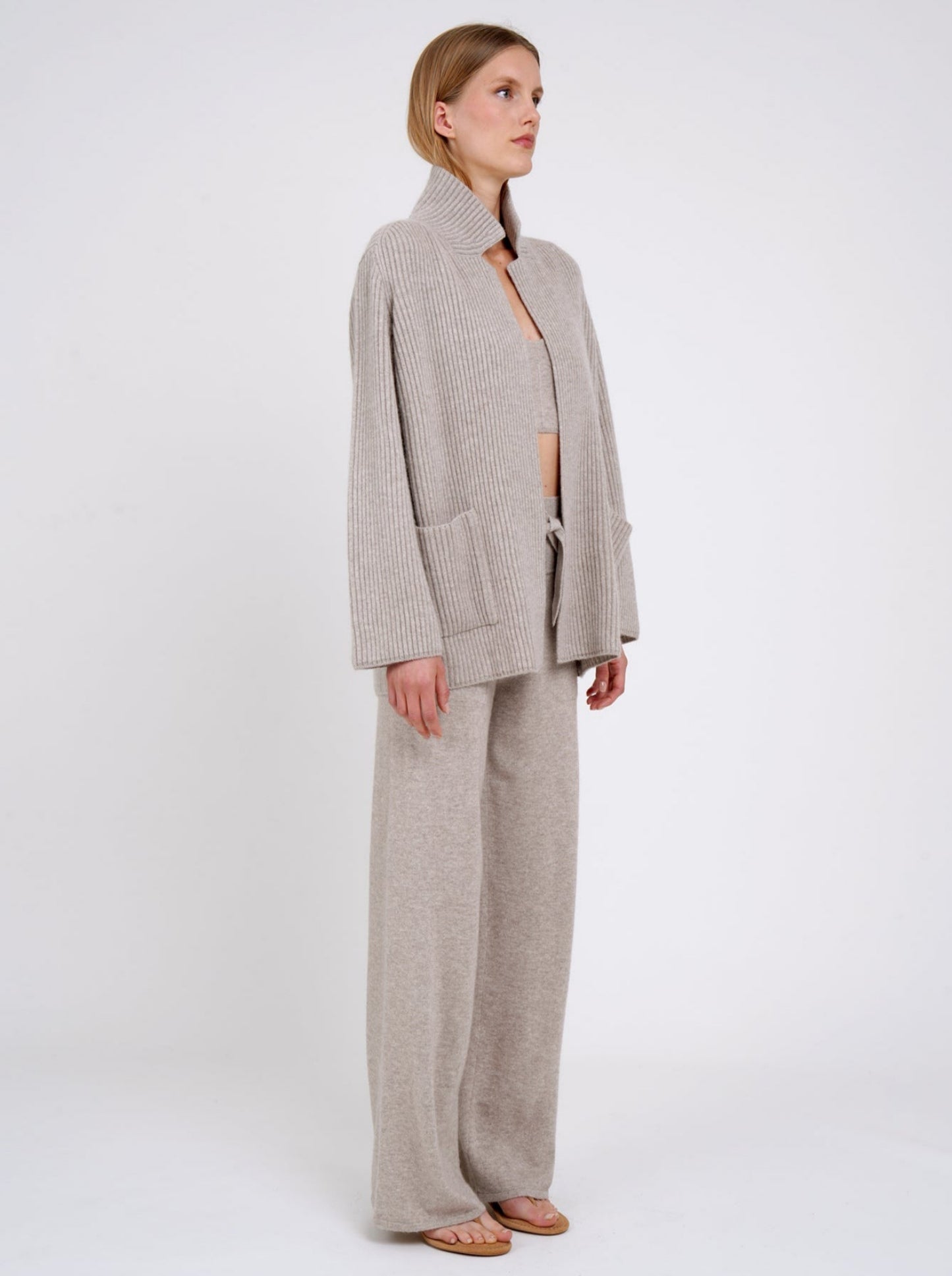 Side view model wearing beige ribbed knit cashmere jacket with high collar and patch pockets; worn with beige cashmere pants.