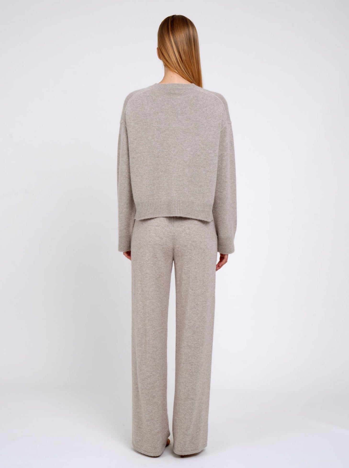Back view model wearing beige cashmere cropped jumper with round neck, long sleeves; worn with matching beige cashmere knit pants.
