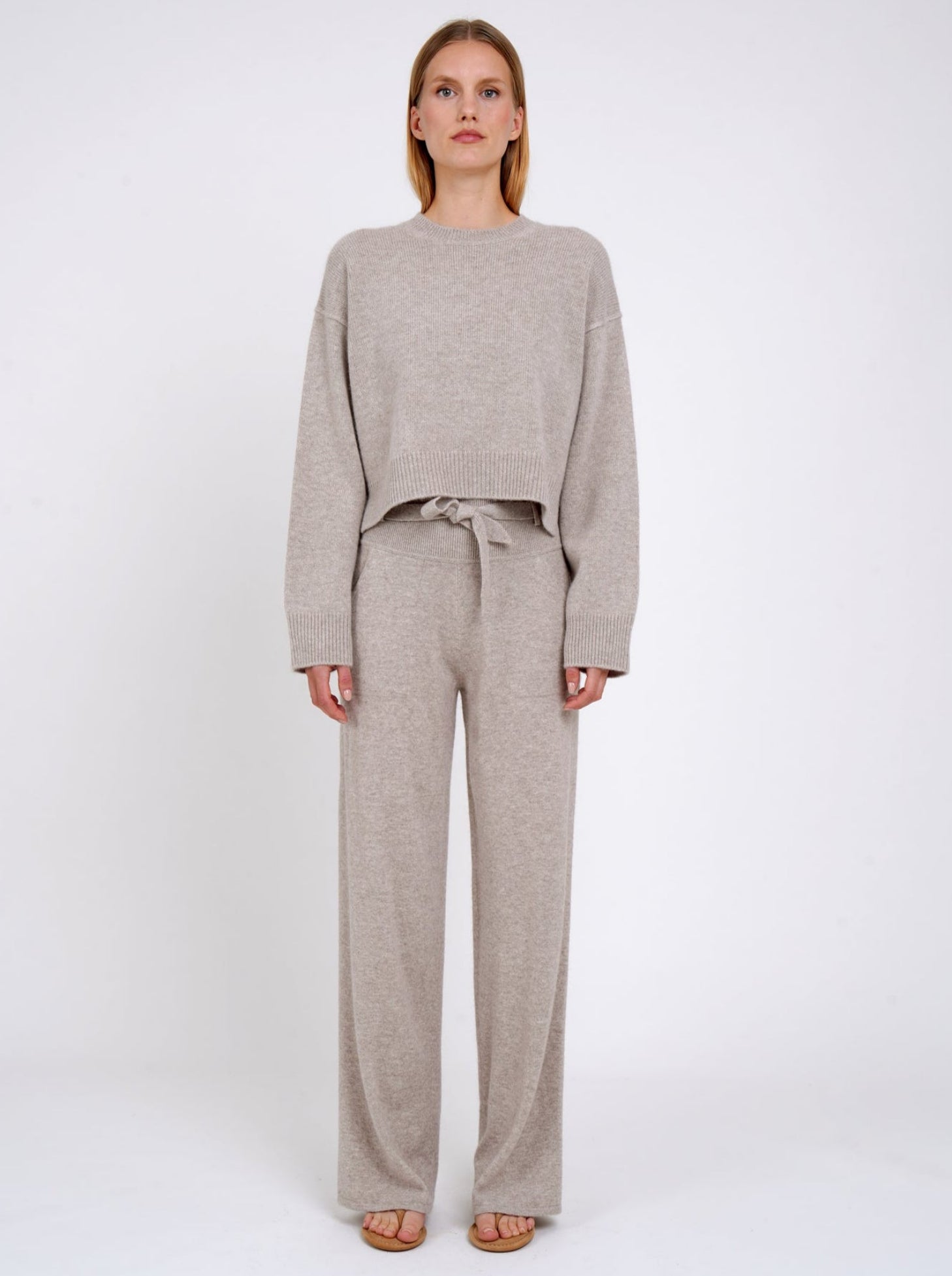 Model wears beige cashmere cropped sweater with round neck, long sleeves; worn with matching beige cashmere, wide leg, belted pants.