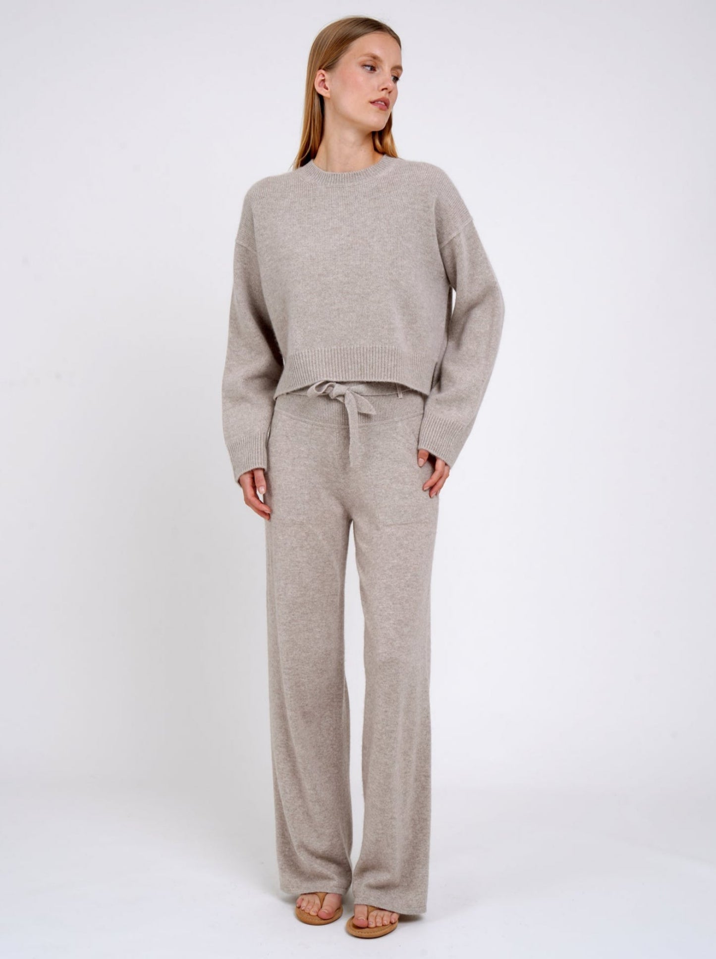 Model wears beige cashmere cropped sweater with round neck, long sleeves; worn with matching beige cashmere, wide leg pants with knitted belt tied at the waist.