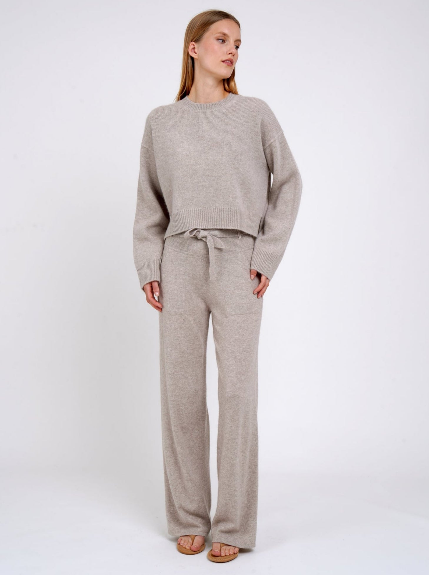 Model wears beige cashmere cropped sweater with round neck, long sleeves; worn with matching beige cashmere, wide leg pants with knitted belt tied at the waist.