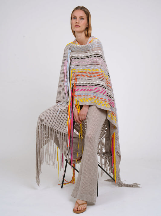 Seated model poses in long beige shawl decorated with hand woven multi-colour fringes and matching beige cashmere knit pants.