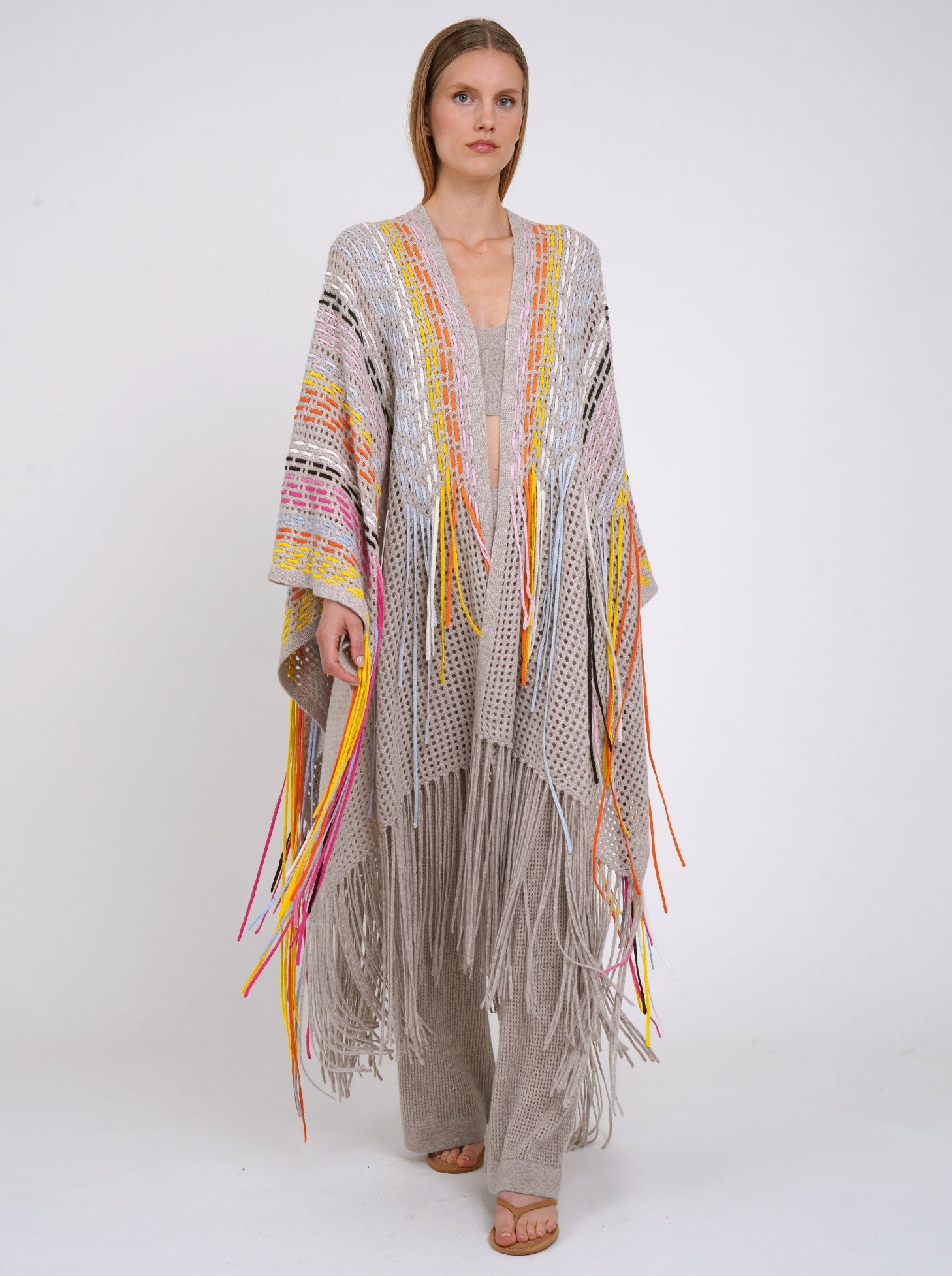 Model walks to camera in long beige shawl decorated with hand woven multi-colour fringes and matching beige cashmere knit pants.