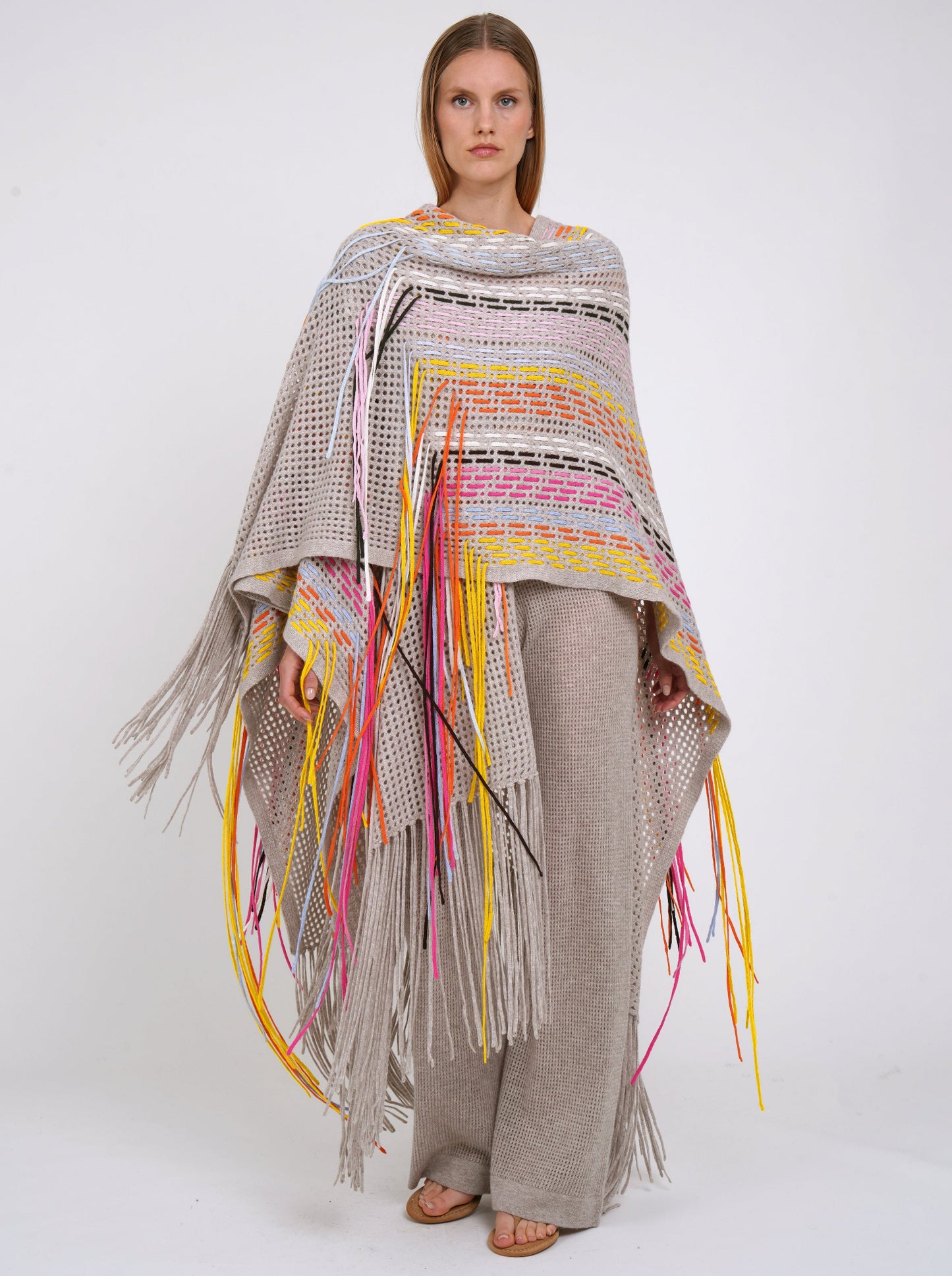 Action shot model in long beige shawl decorated with hand woven multi-colour fringes and matching beige cashmere knit pants.