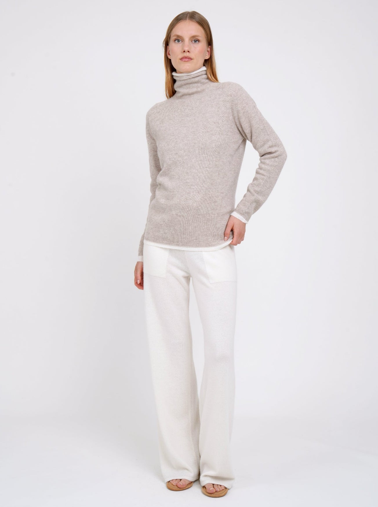 Woman wearing a beige turtle neck sweater and white pants on a white background.