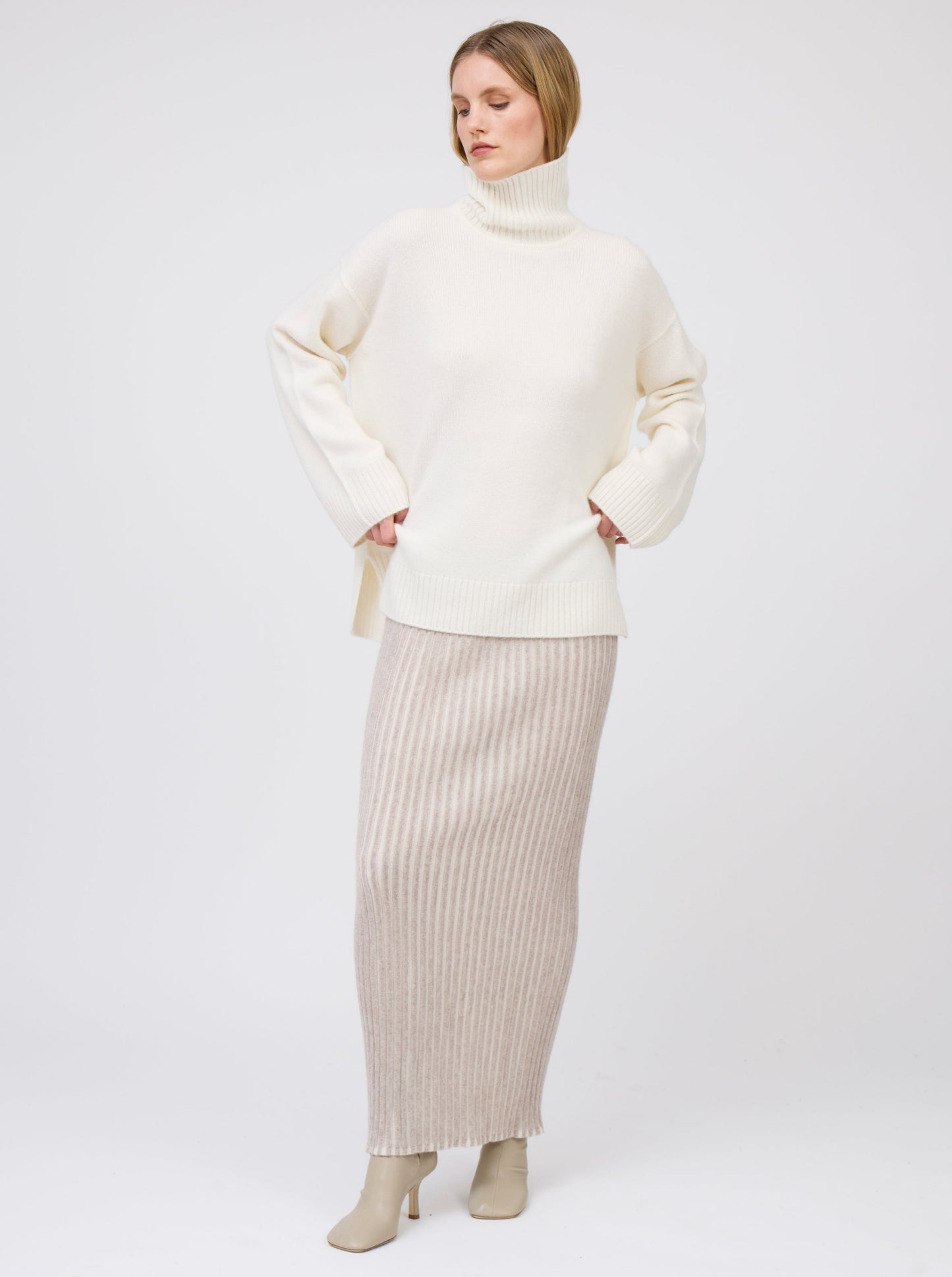 Front view model wears maxi length slim tube skirt in beige and white vertical stripes; worn with oversized natural white roll neck cashmere jumper and beige heeled boots.