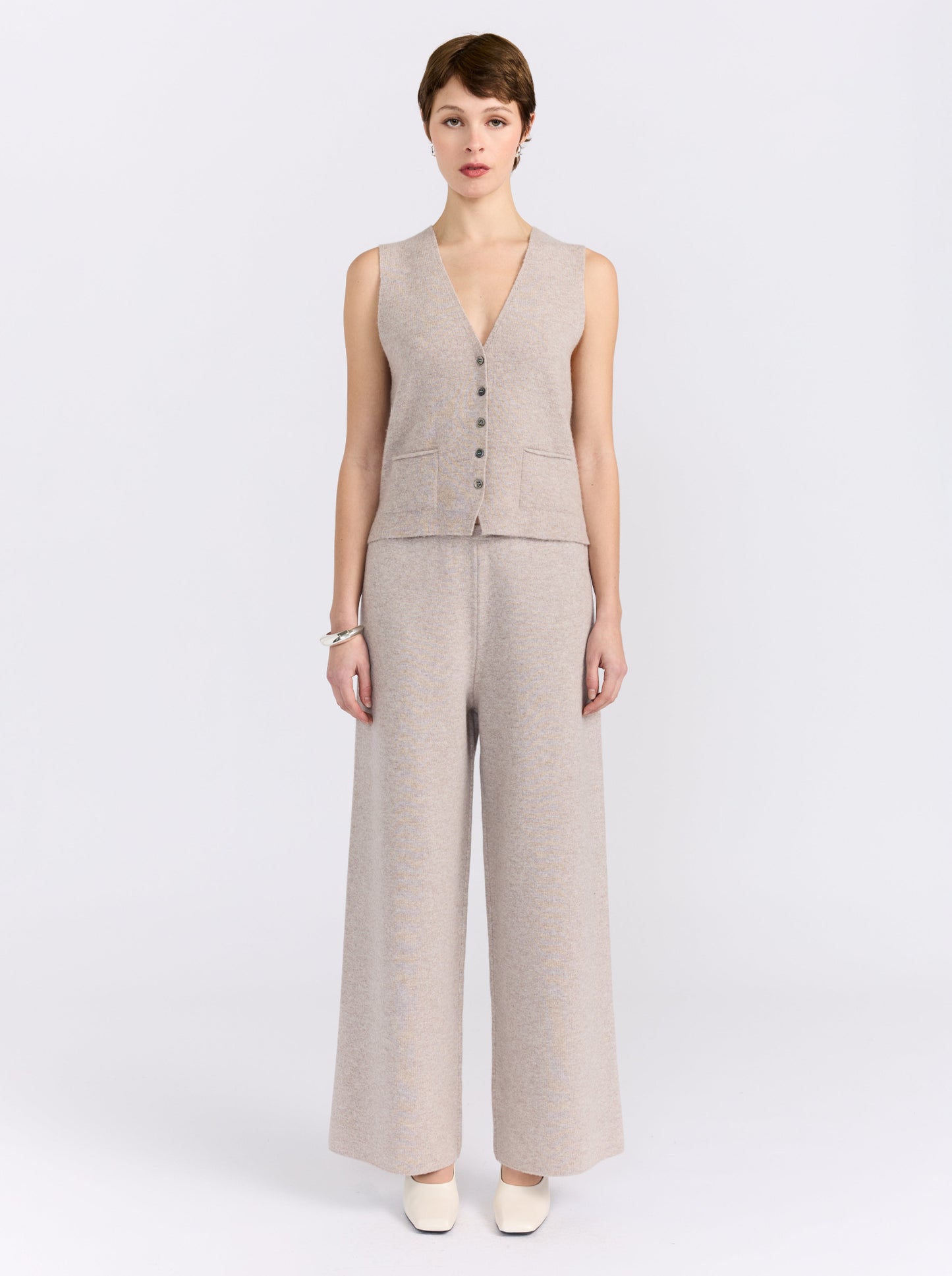 A model stands against a light gray studio background wearing a sleeveless beige knit vest with front buttons and matching wide-leg trousers.