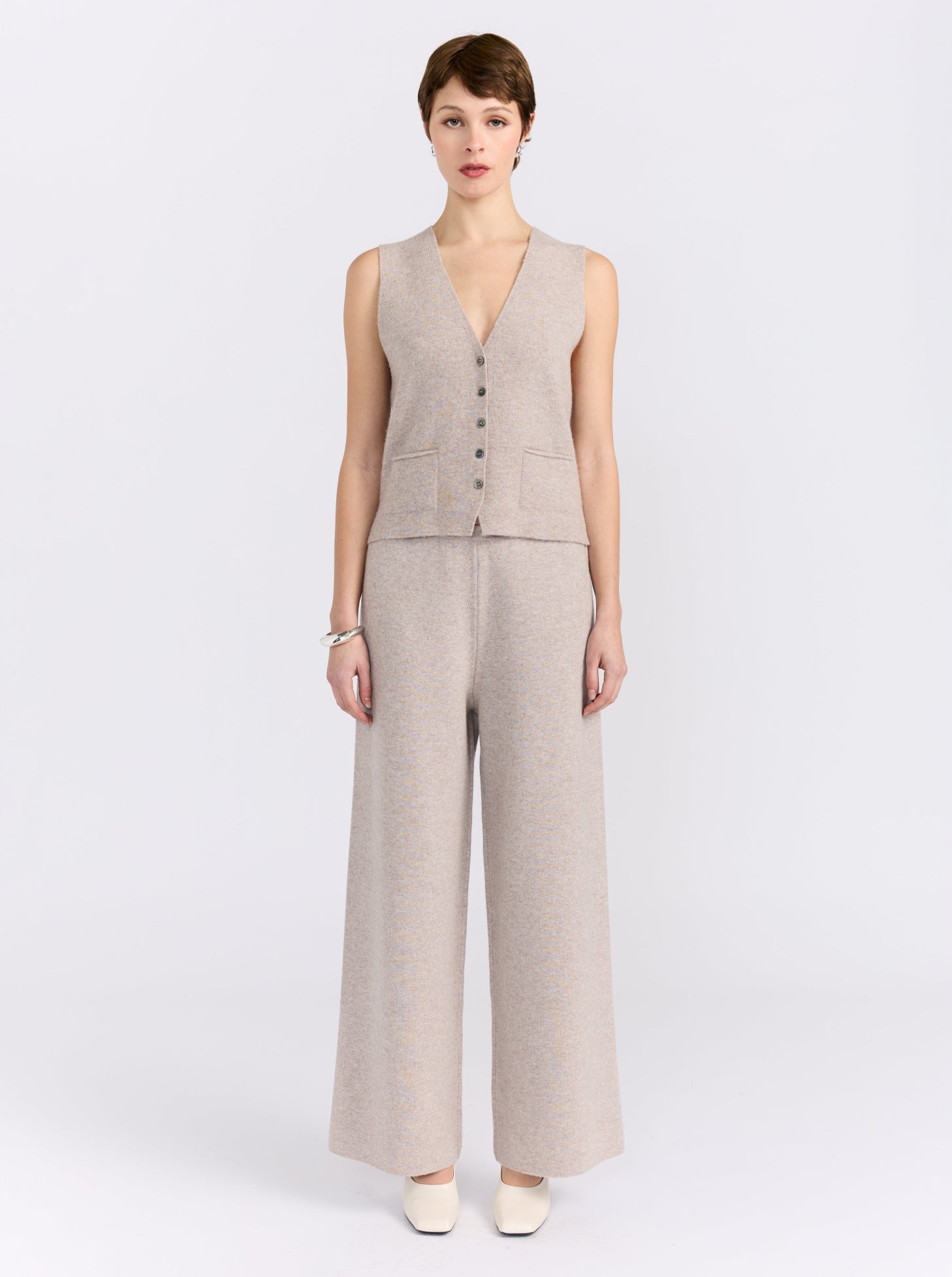 A model stands against a light gray studio background wearing a sleeveless beige knit vest with front buttons and matching wide-leg trousers.