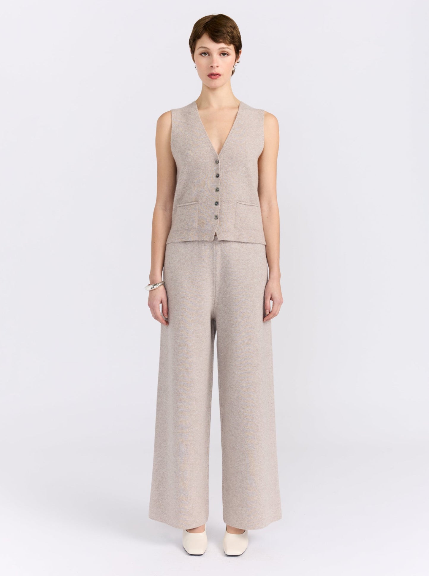 A model stands against a light gray studio background wearing a sleeveless beige knit vest with front buttons and matching wide-leg trousers.