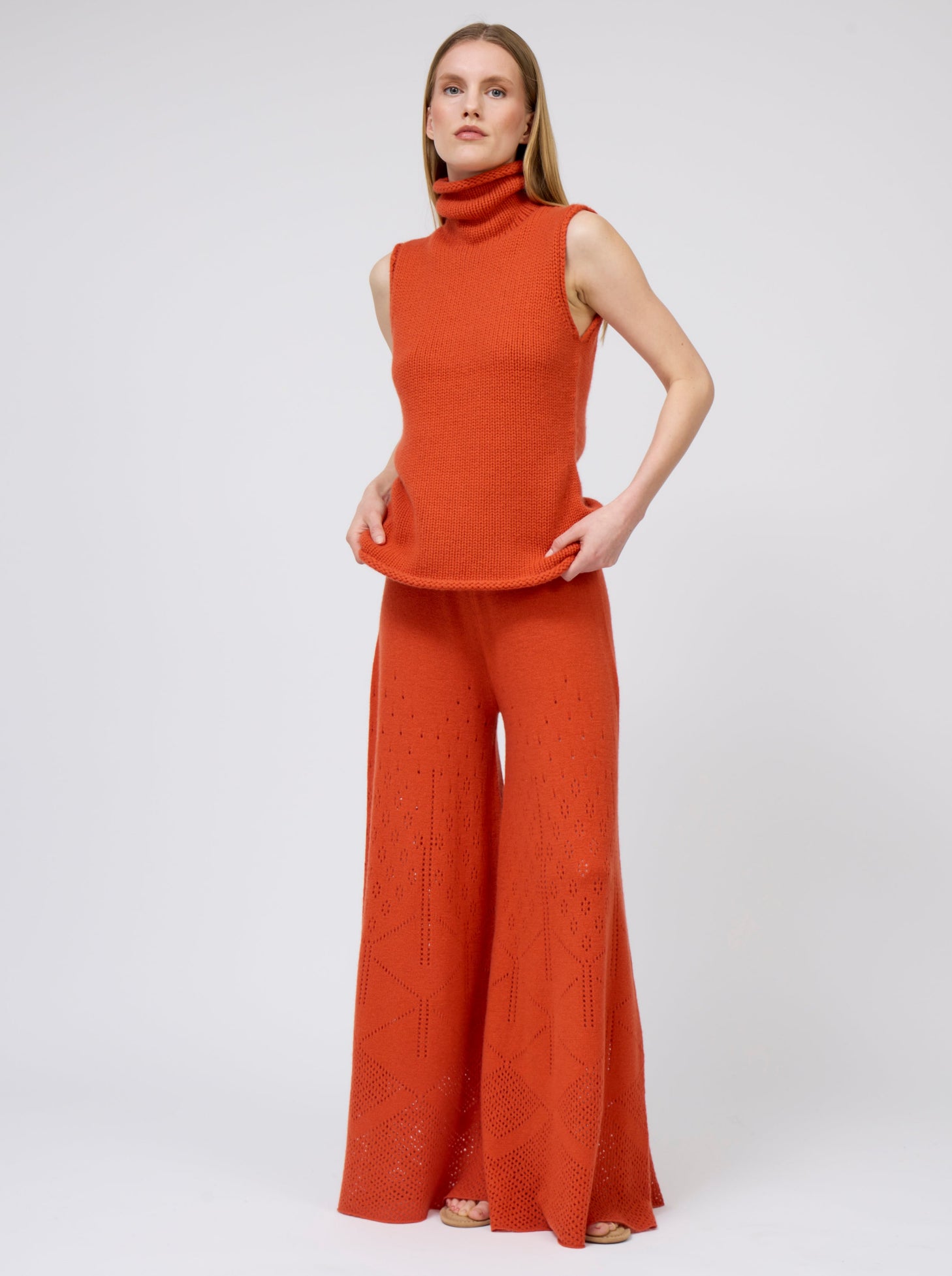 Front view model wears bright orange sleeveless chunky cashmere knit roll neck jumper with matching orange pants.