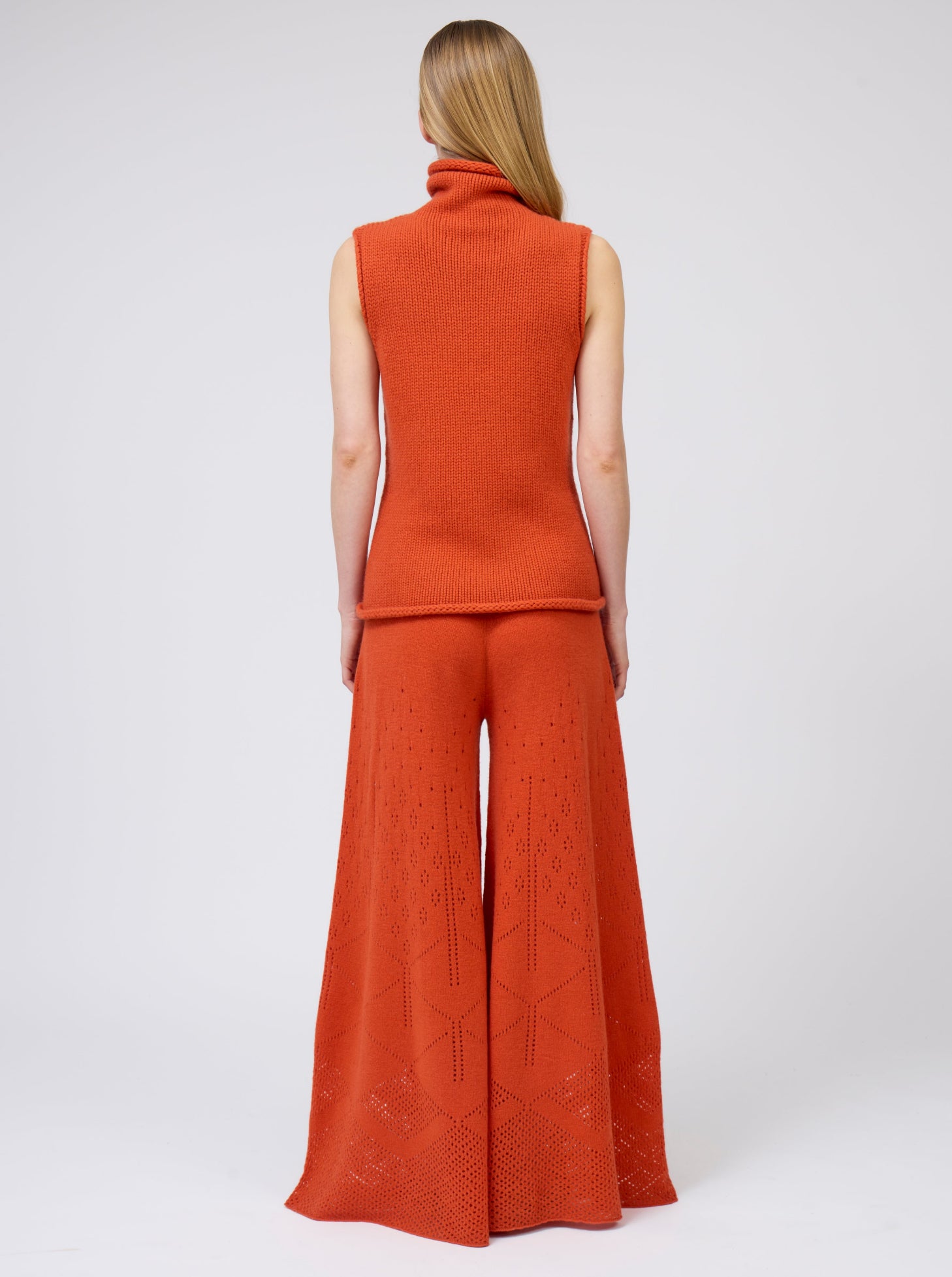 Back view model wears bright orange sleeveless chunky cashmere knit roll neck jumper with very flared cut, matching orange pants.
