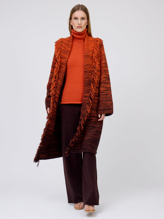 Model walks towards viewer wearing orange/black mix knit cashmere coat with fringe trim shawl collar; worn with orange jumper and brown pants.