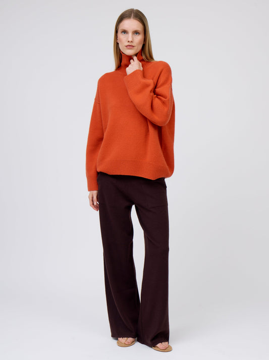 Posed model wears deep orange cashmere high roll neck jumper with wide leg dark brown pants and suede thongs.