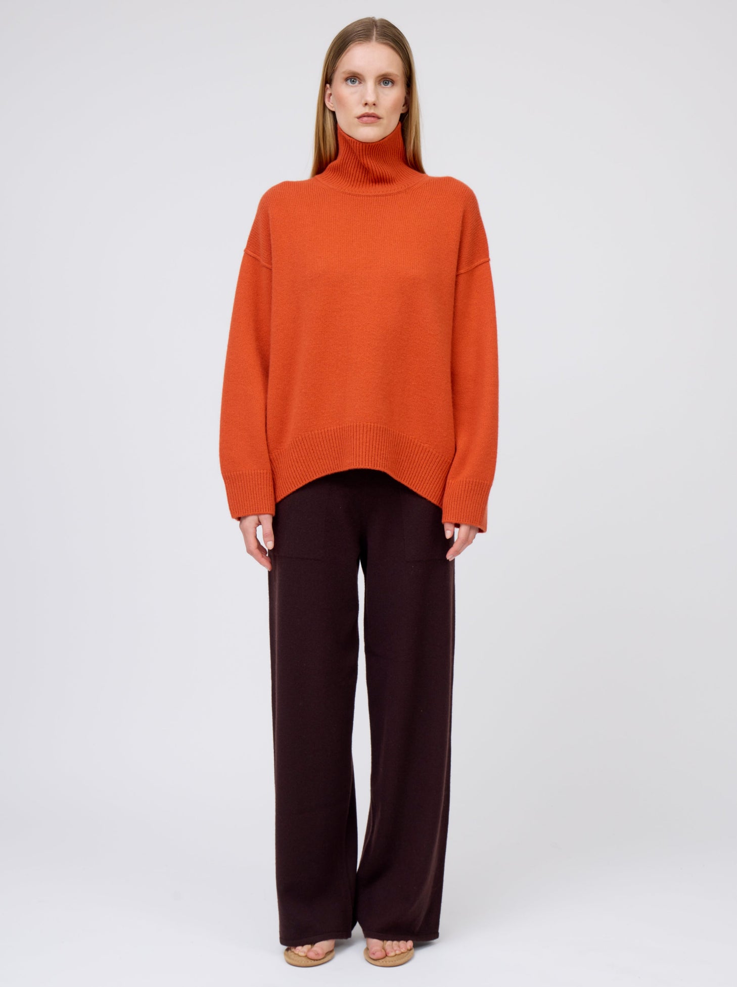 Straight front view model wears deep orange cashmere roll neck jumper with wide leg dark brown pants.