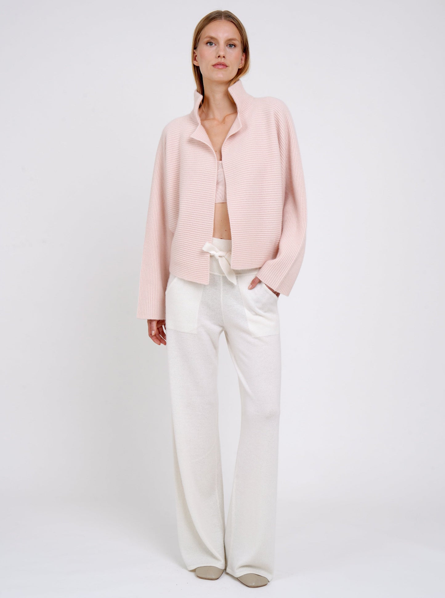 Model wears relaxed fit peach ribbed knit cashmere jacket with high collar kimono sleeves and white wide leg knit pants.