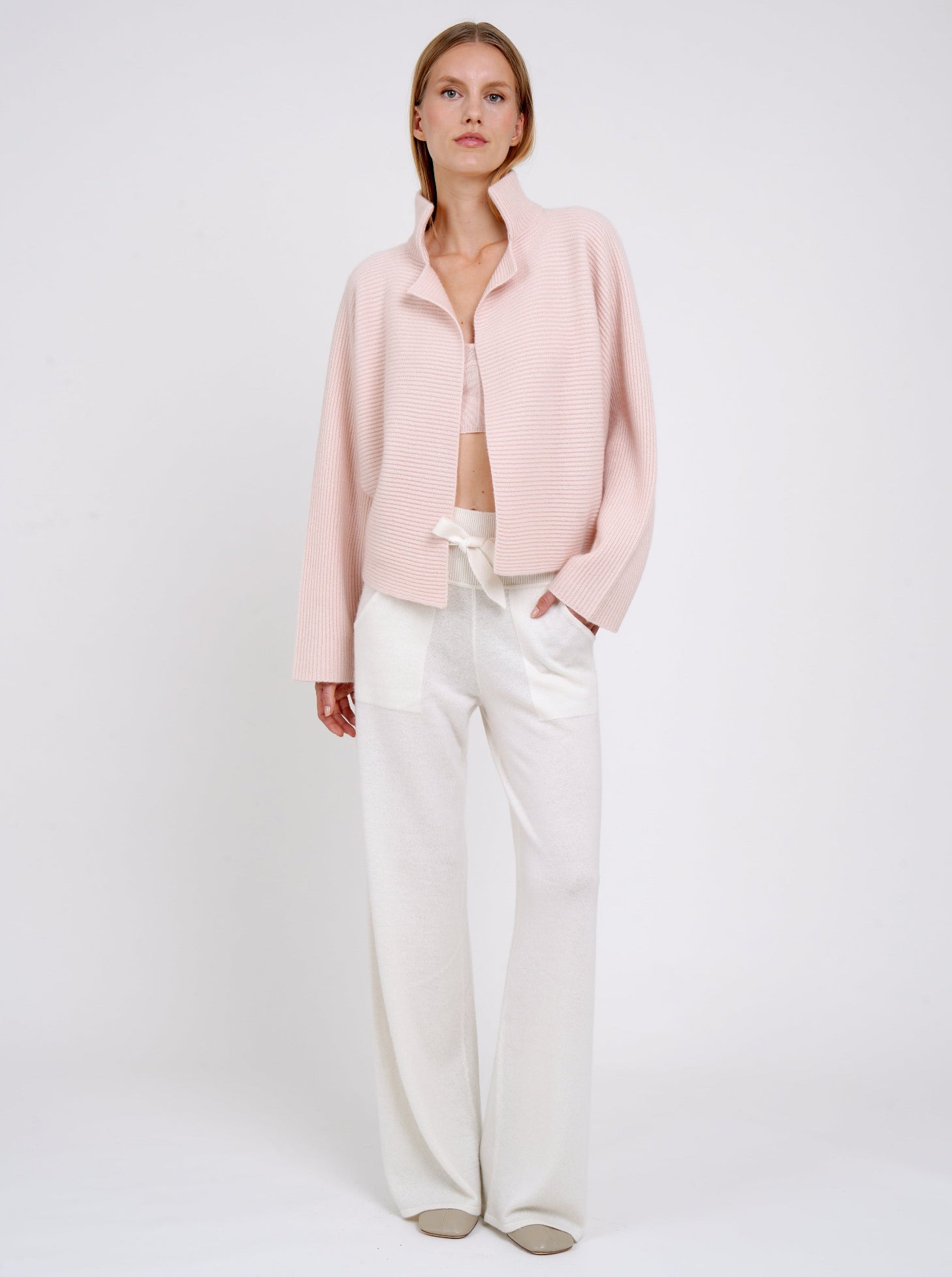 Model wears relaxed fit peach ribbed knit cashmere jacket with high collar kimono sleeves and white wide leg knit pants.
