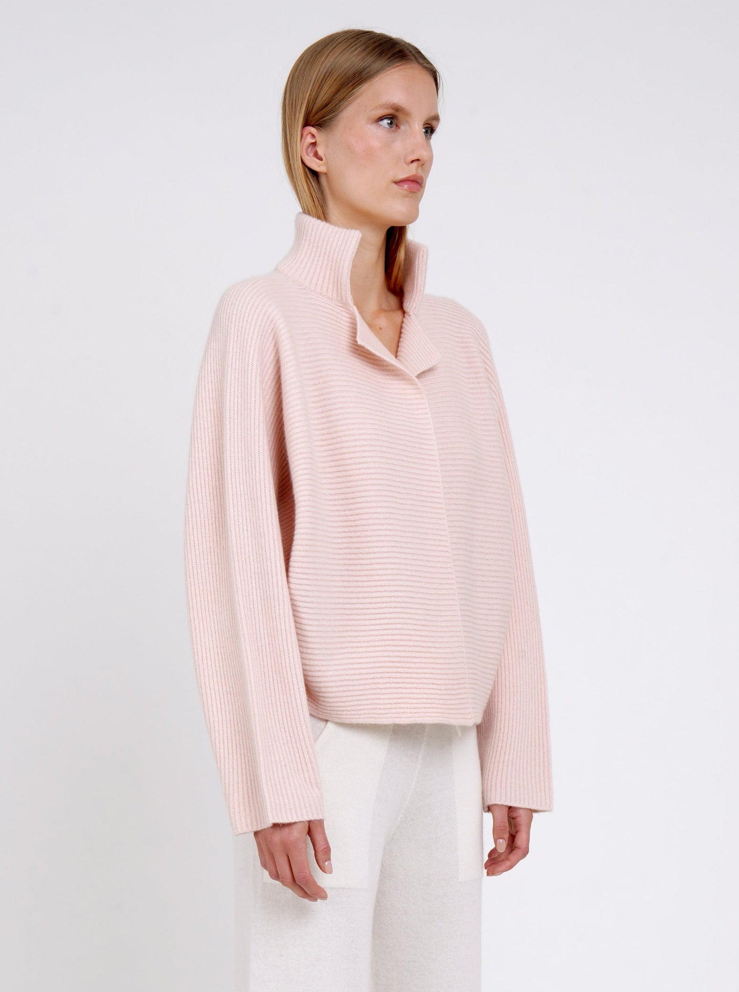 Side view close-up modal in relaxed fit ribbed knit peach cashmere jacket with high collar kimono sleeves and white wide leg knit pants.
